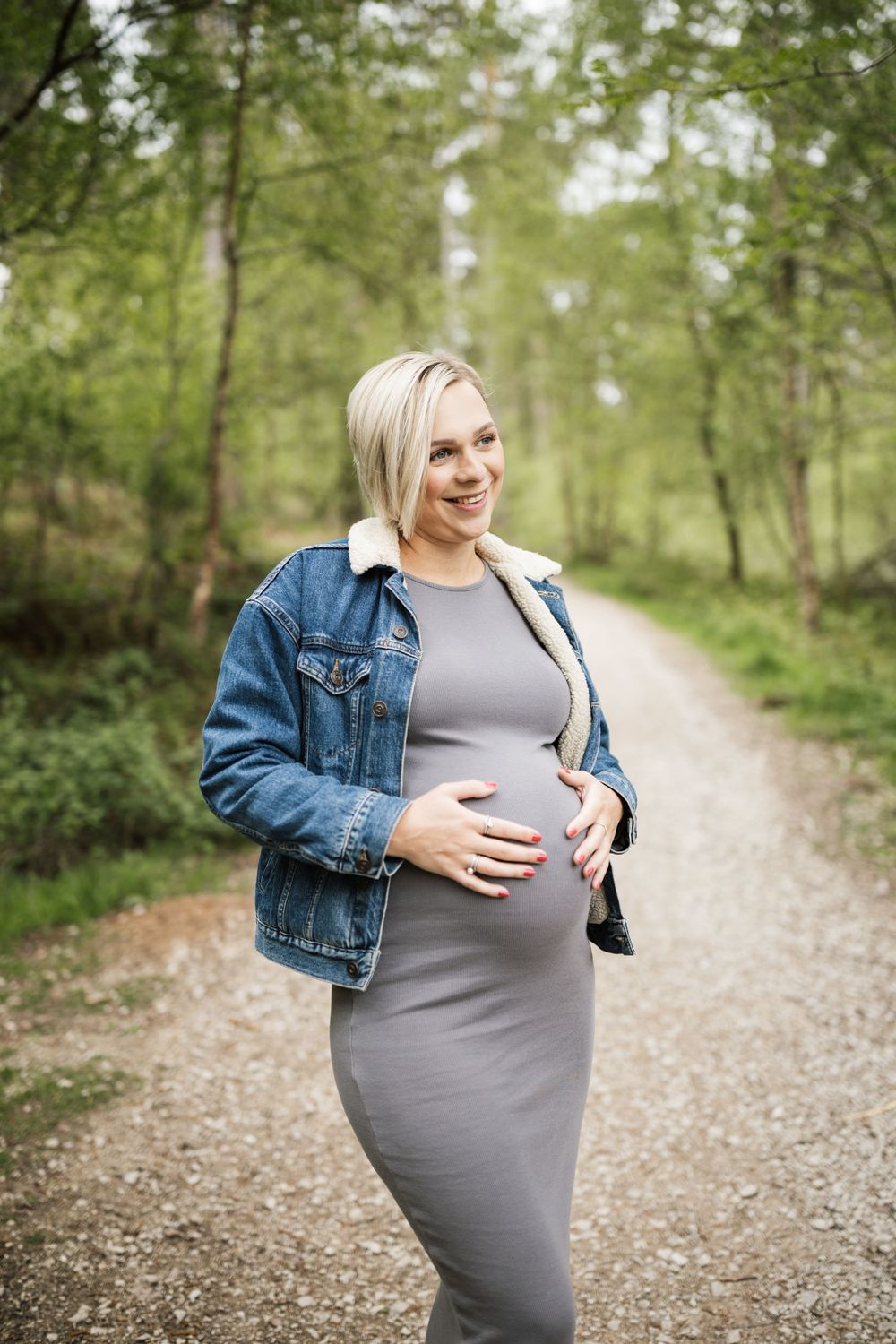 A person in a denim jacket and gray dress walks along a serene gravel path surrounded by lush green trees.