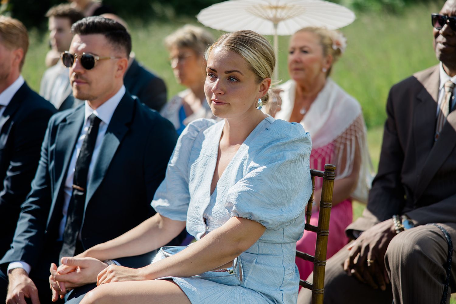 Wedding guests seated outdoors in rows of white chairs on a sunny day with a decorative white parasol visible.