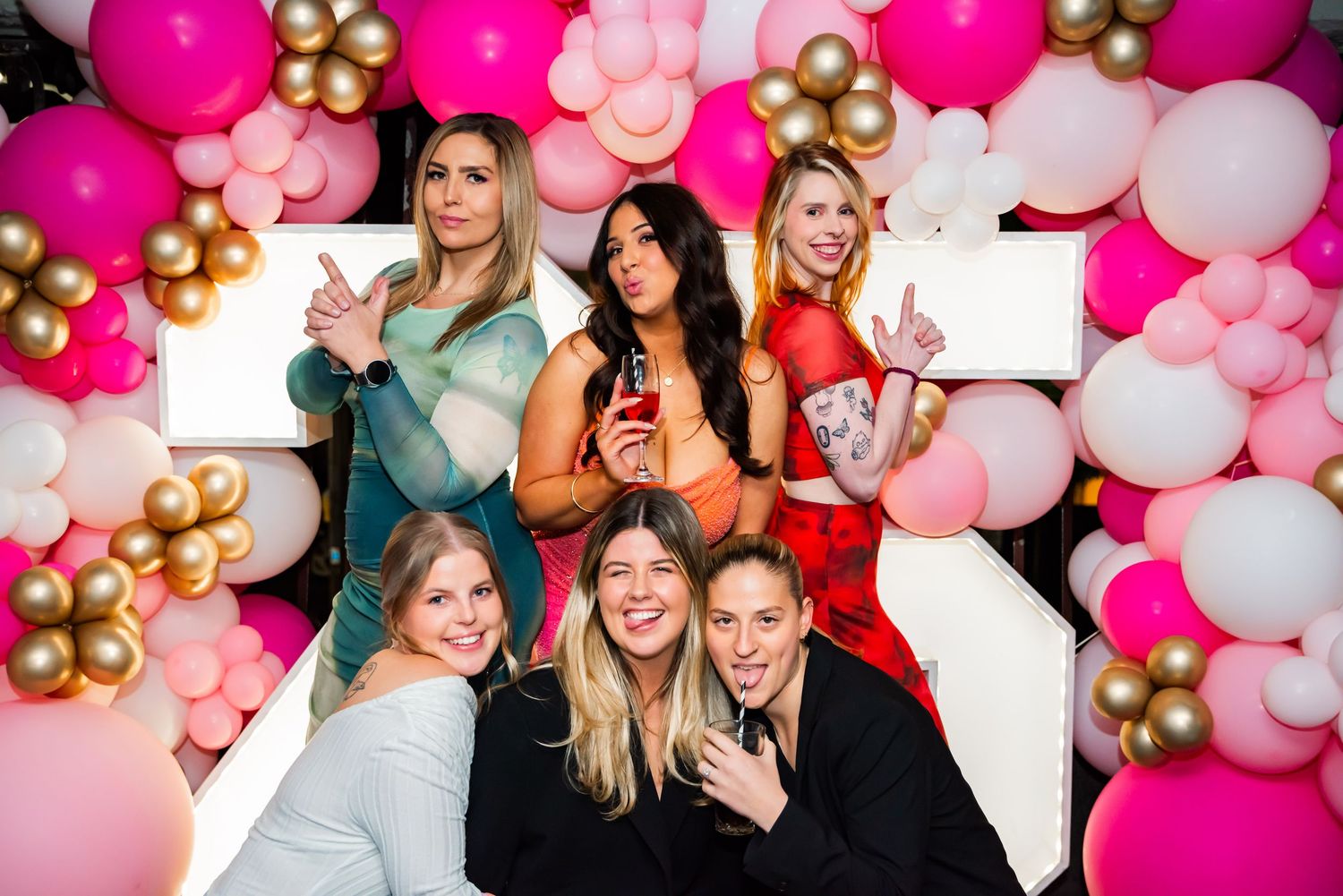 Friends laugh and celebrate together against a festive backdrop of pink, white and gold balloons at a party.