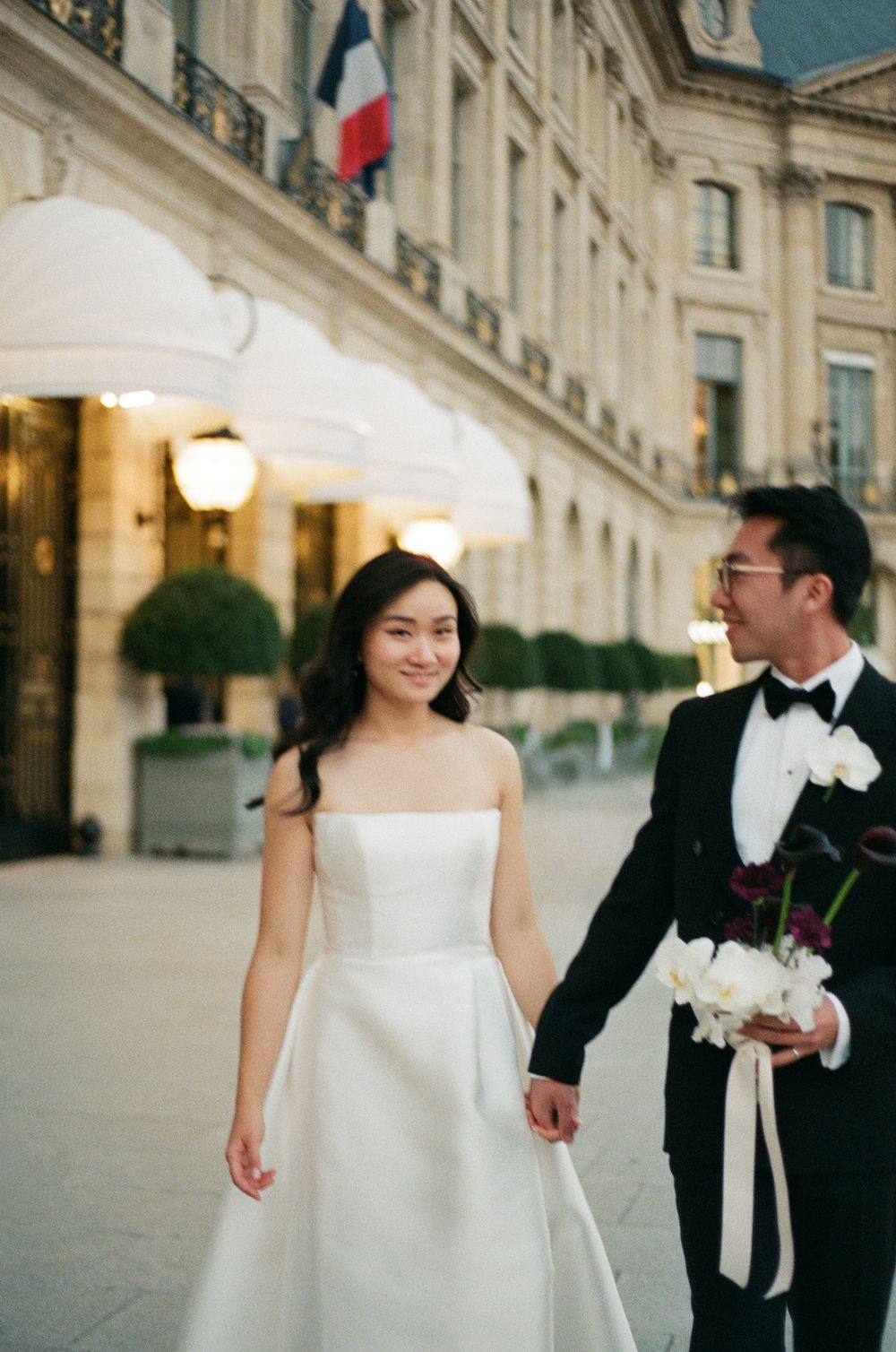 Couple in wedding attire holding hands outside a Parisian building, bride in strapless white gown,. YSL suit. Ritz paris wedding photography