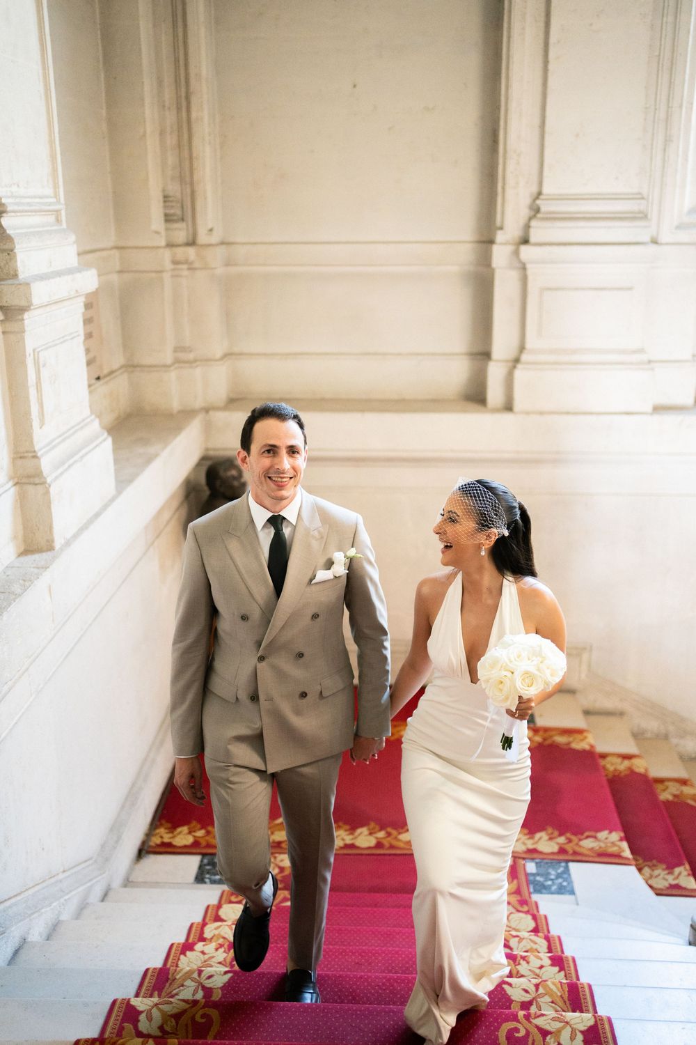Bride and groom walk up red-carpeted stairs holding hands, she in white halter gown. Paris mairie wedding, paris civil wedding, film photo