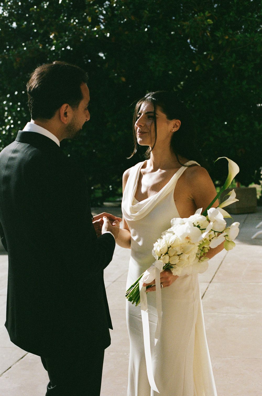 Bride in white cowl-neck gown holding white calla lily bouquet exchanges vows with groom in black tuxedo outdoors. Paris intimate wedding