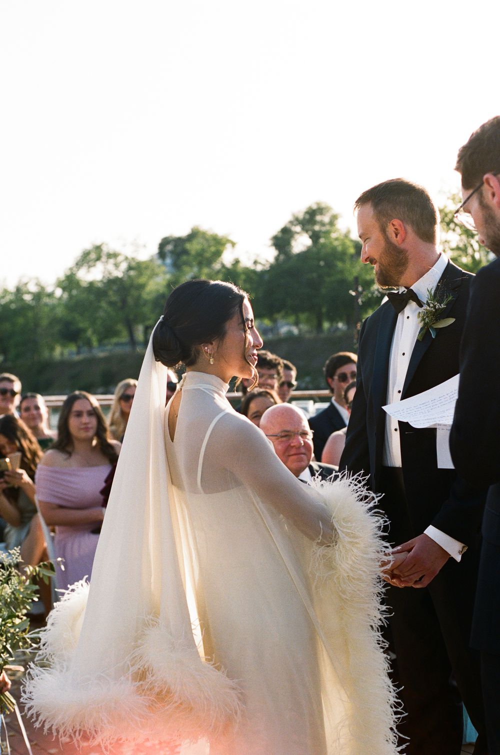 Bride in feather-trimmed cape wedding dress holds hands with groom in tuxedo during outdoor ceremony.