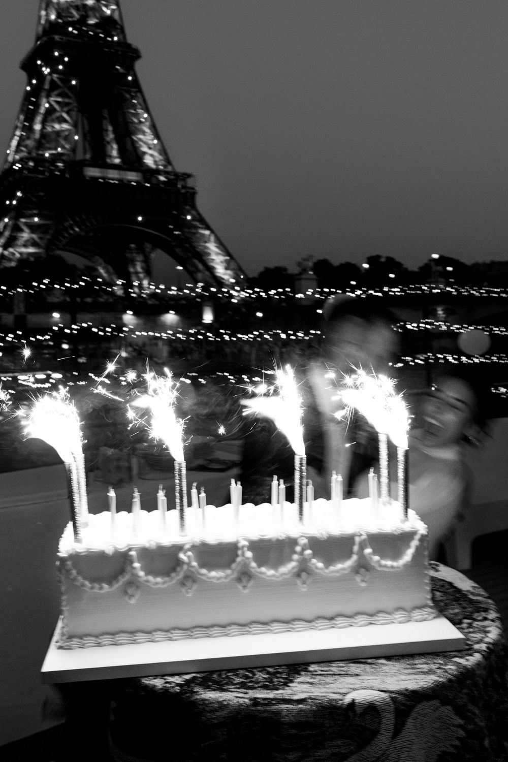 Black and white photo of a lit birthday cake with sparklers in front of the illuminated Eiffel Tower at night in Paris.