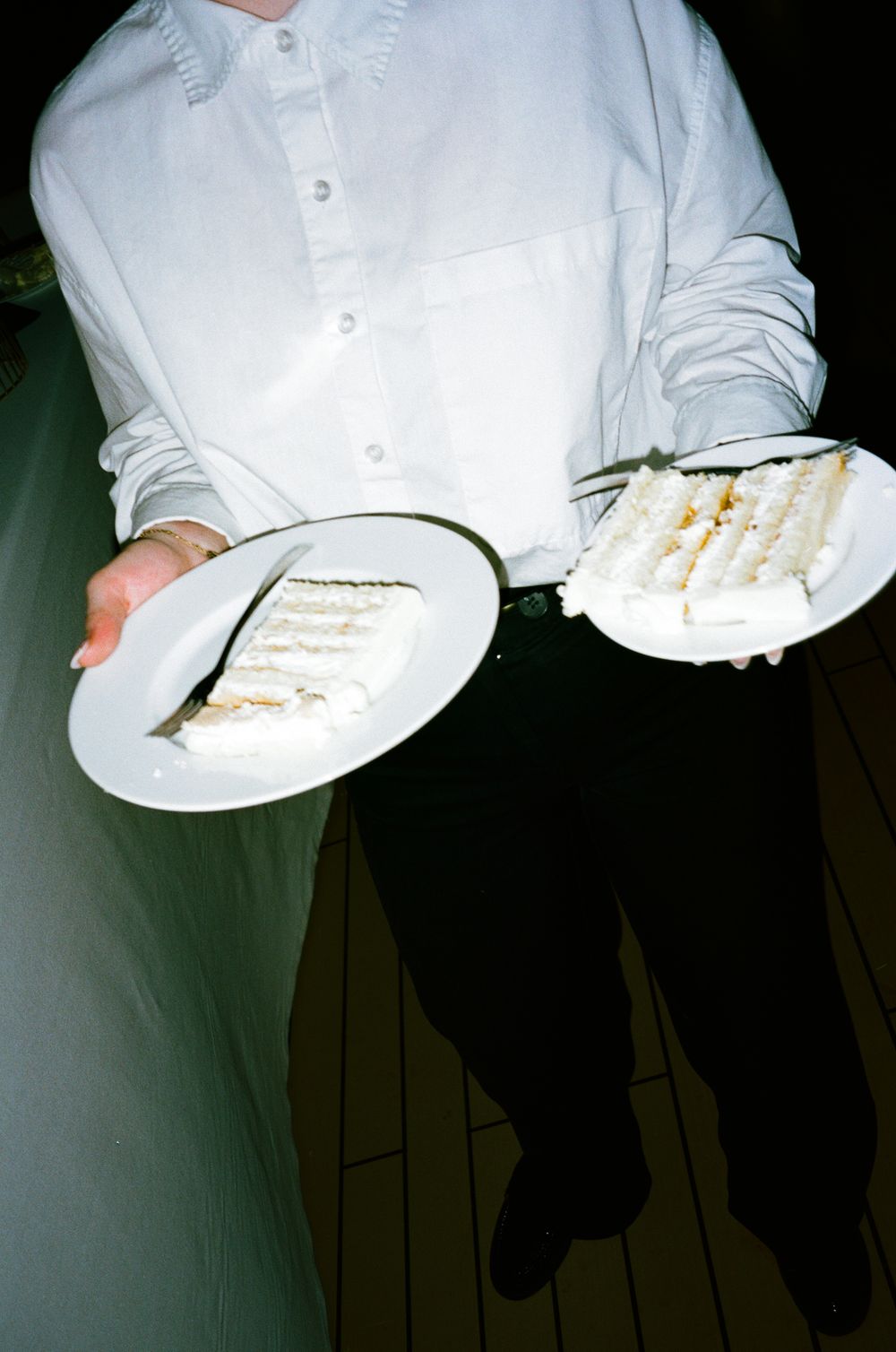 Person in white shirt holding two white plates with slices of white frosted cake at an event.