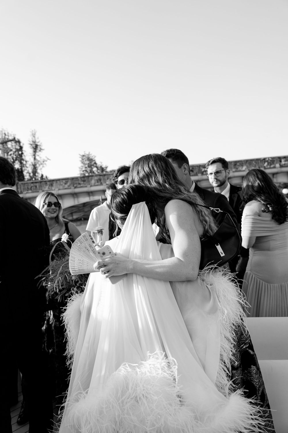 Black and white photo of a bride in a feathered gown embracing a guest at an outdoor wedding reception.