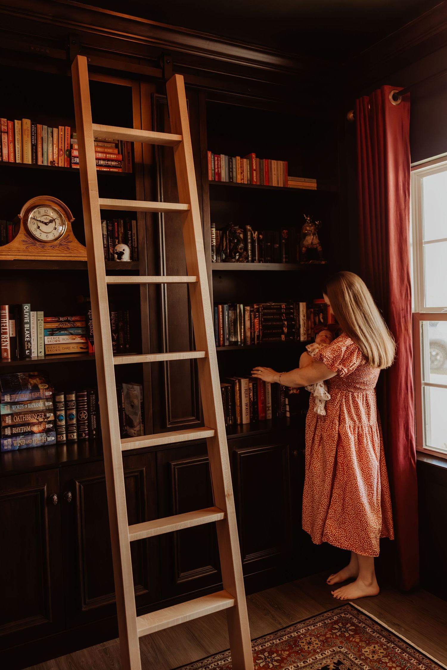 A wooden library ladder leans against dark bookshelves while someone in a coral dress browses books by a window.