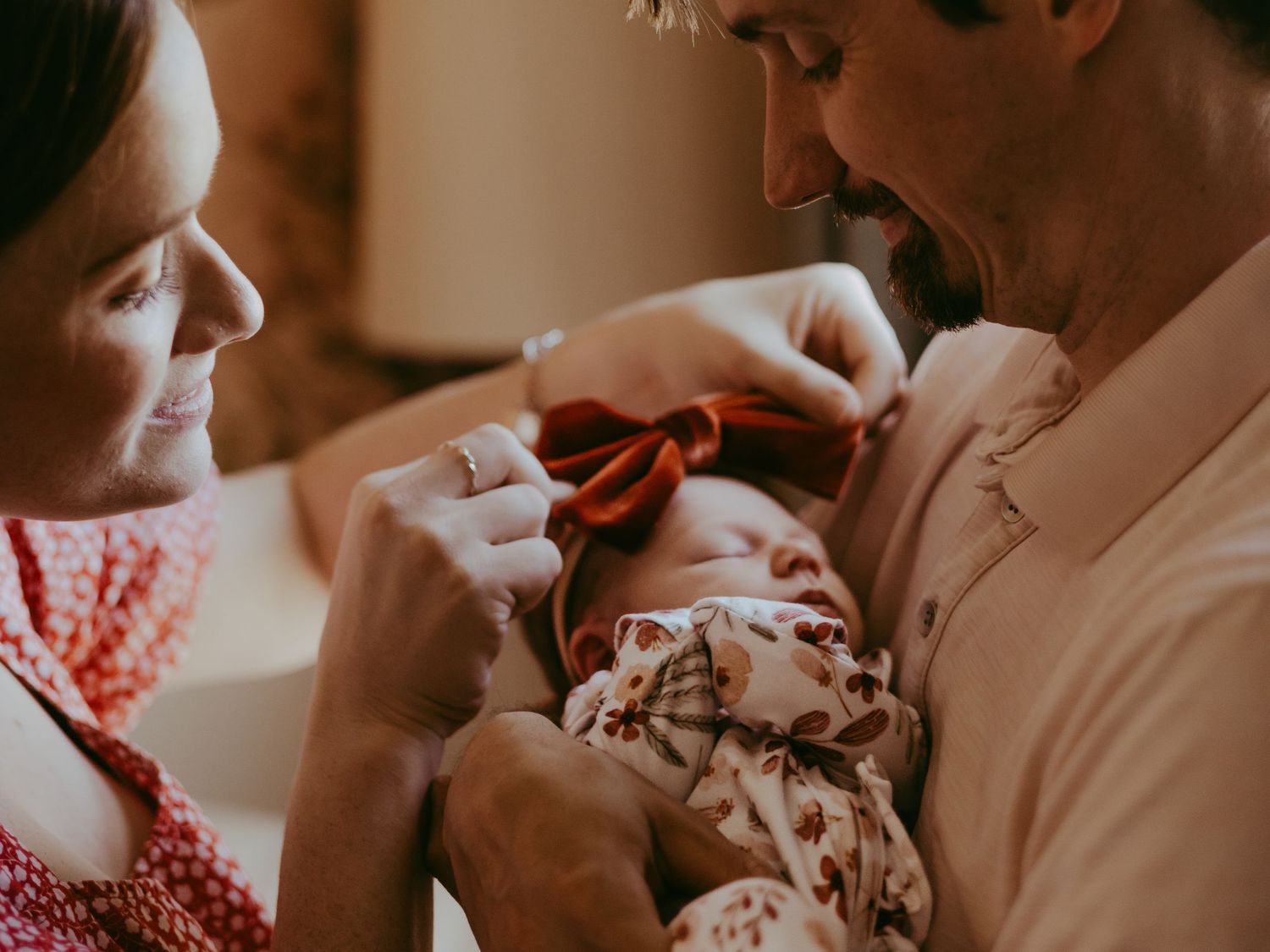 Parents lovingly gaze at their newborn baby wearing a floral outfit in warm, intimate lighting.