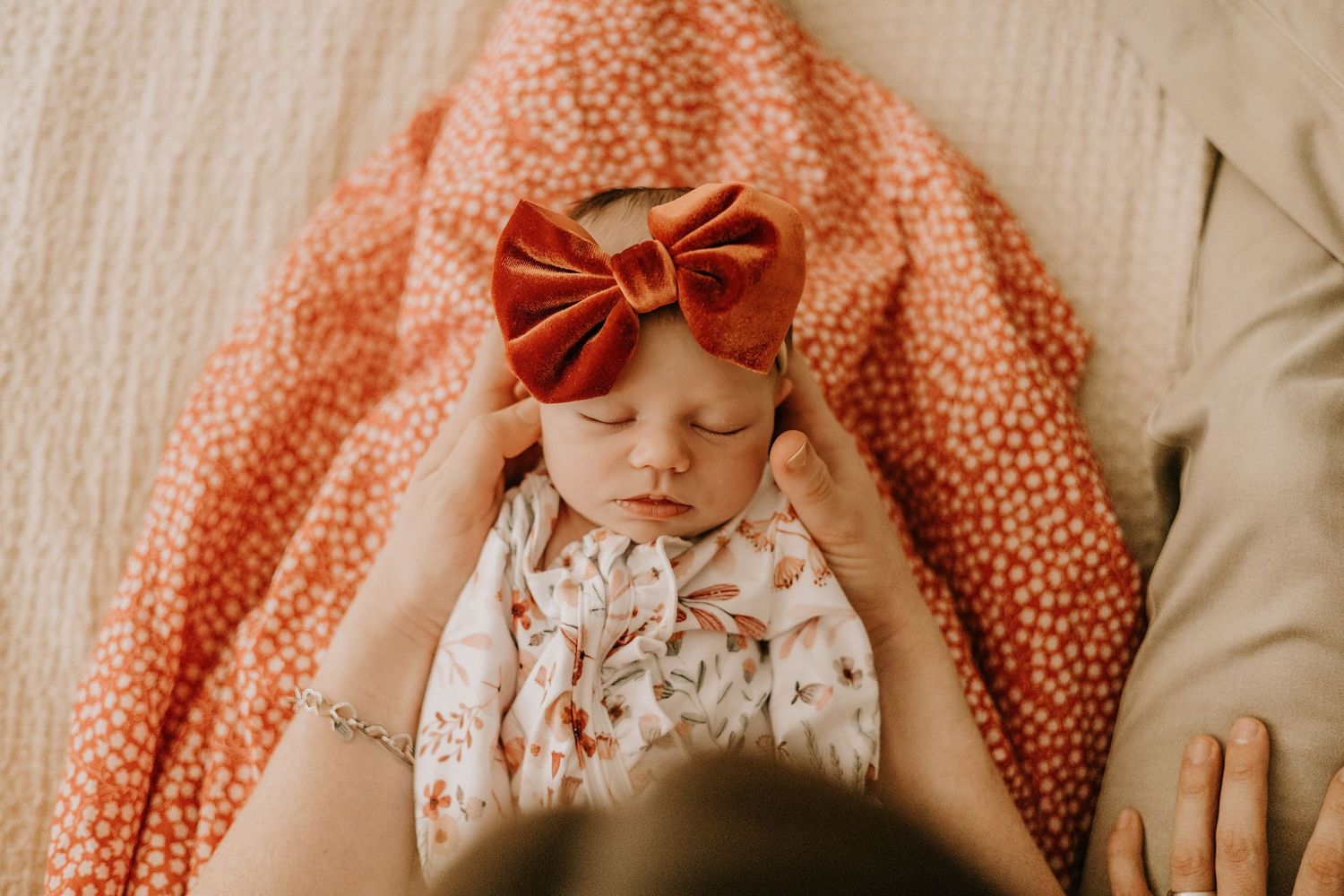 A newborn baby wearing a floral outfit and coral bow headband lies on cream colored bedding.
