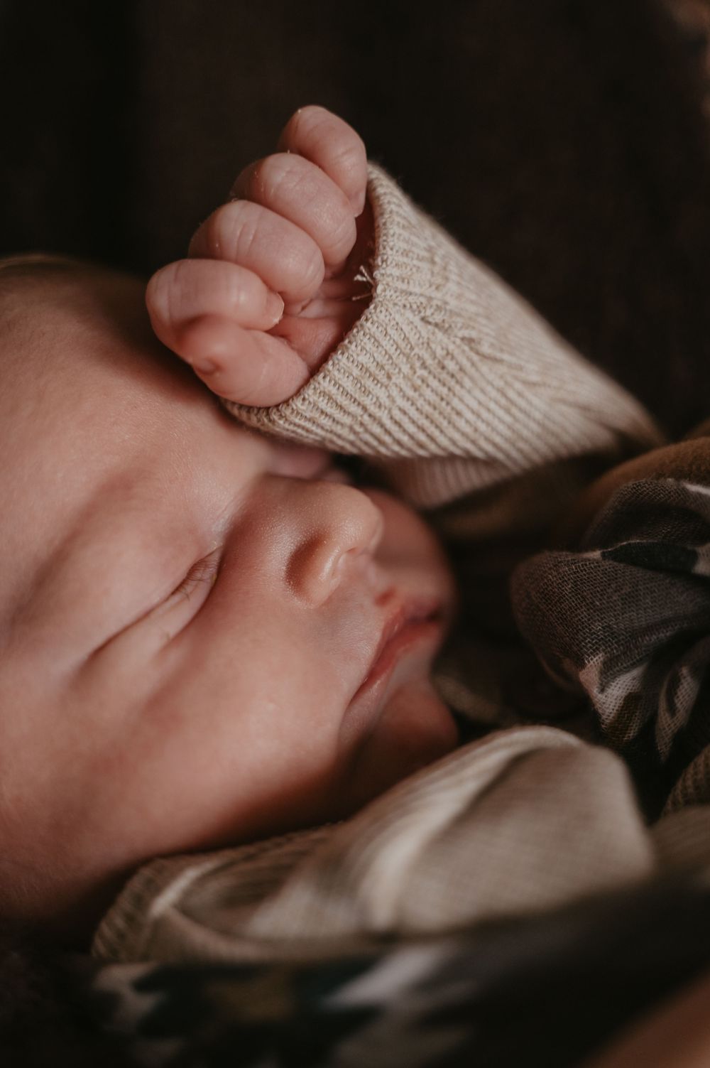 Close-up view of a sleeping baby's face and tiny hand grasping a knit blanket in soft lighting.
