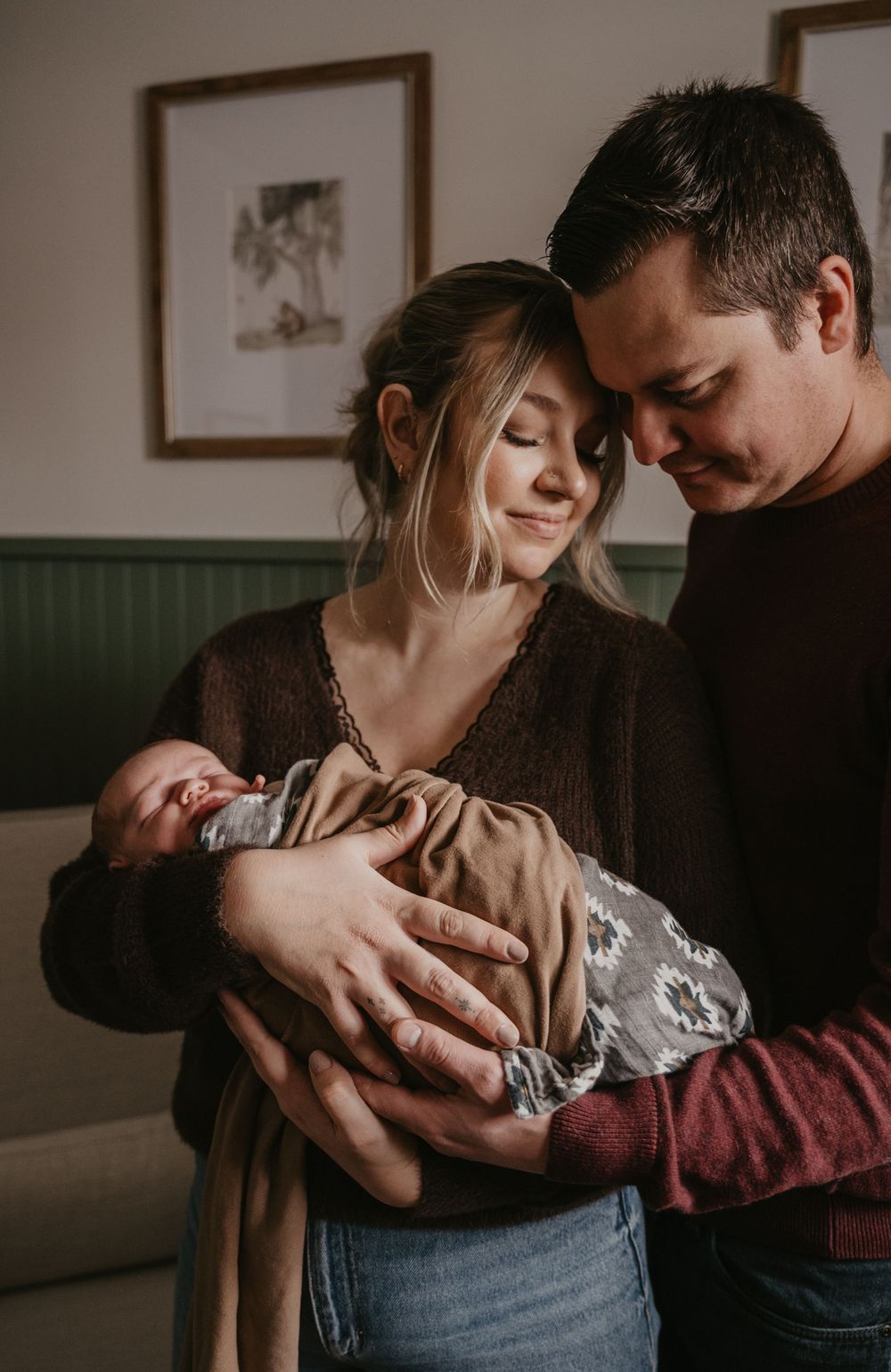 A couple shares an intimate moment while holding their newborn baby in a cozy indoor setting.