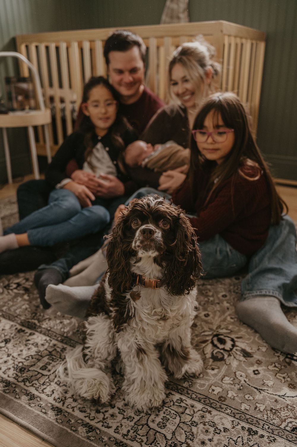 Family sitting on floor with spaniel dog in nursery room with wooden crib in background.