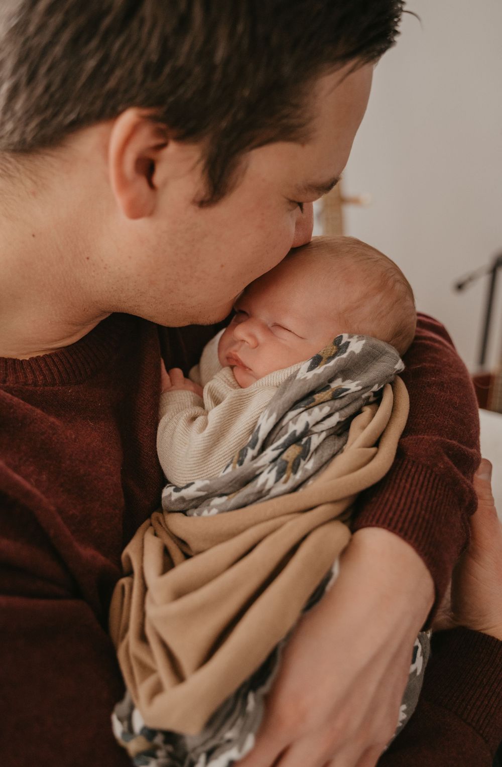 Parent sharing an intimate moment with newborn baby wrapped in beige swaddle against neutral background.