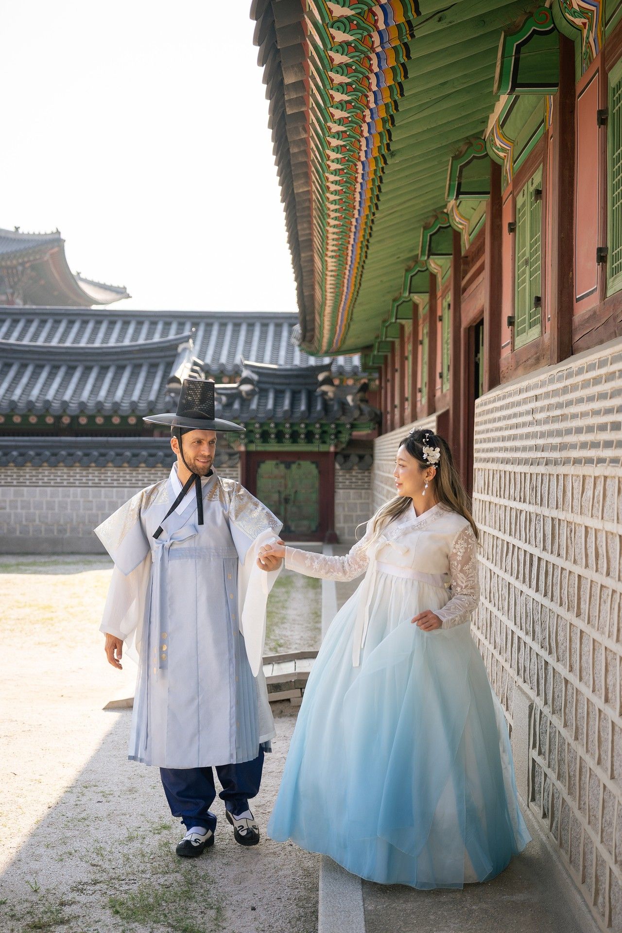A couple in traditional Korean hanbok attire walking along an ornate palace corridor with decorative painted beams.