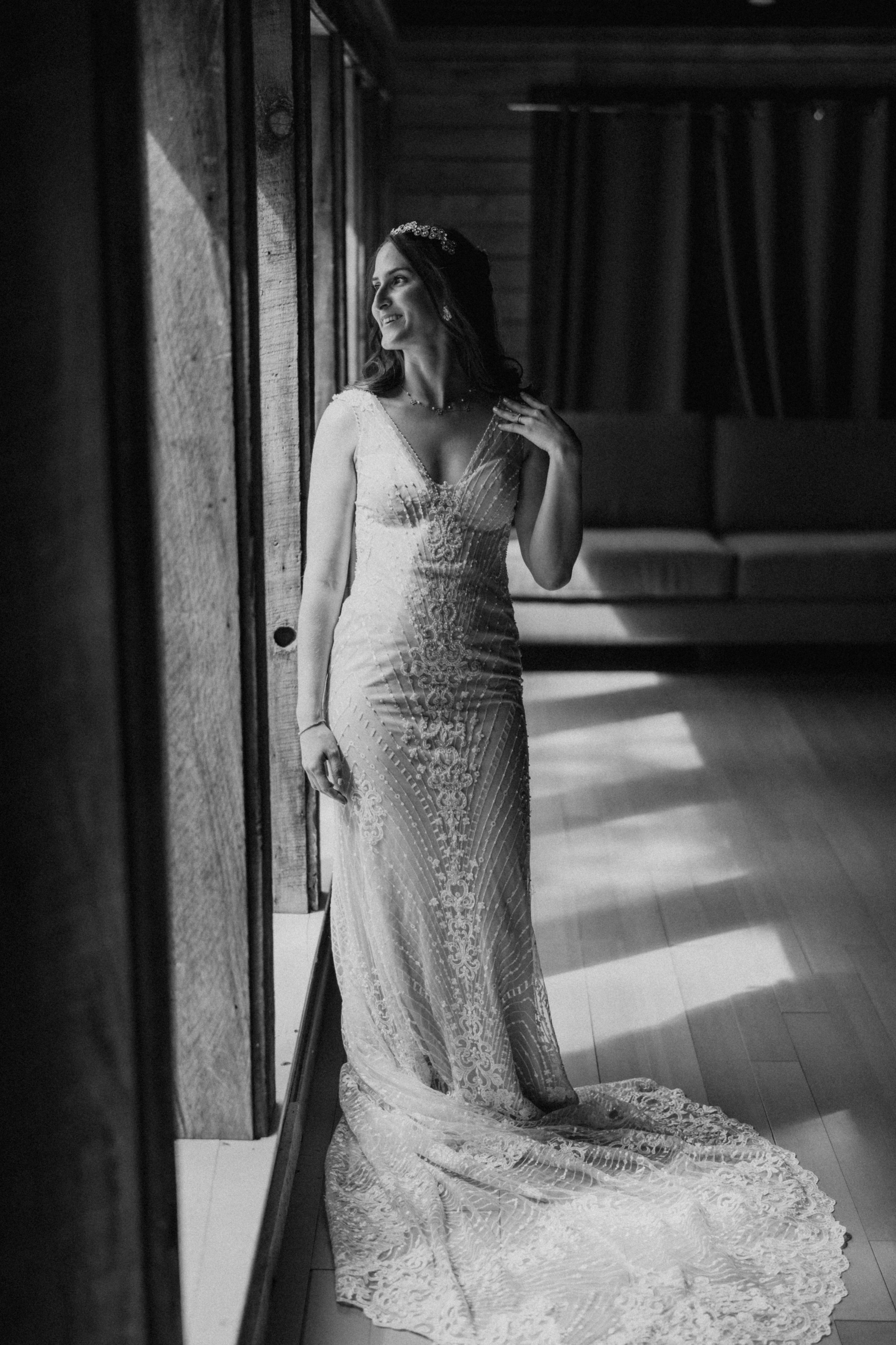 Black and white photo of a bride in a fitted lace wedding gown with long train standing by a window in dramatic lighting.