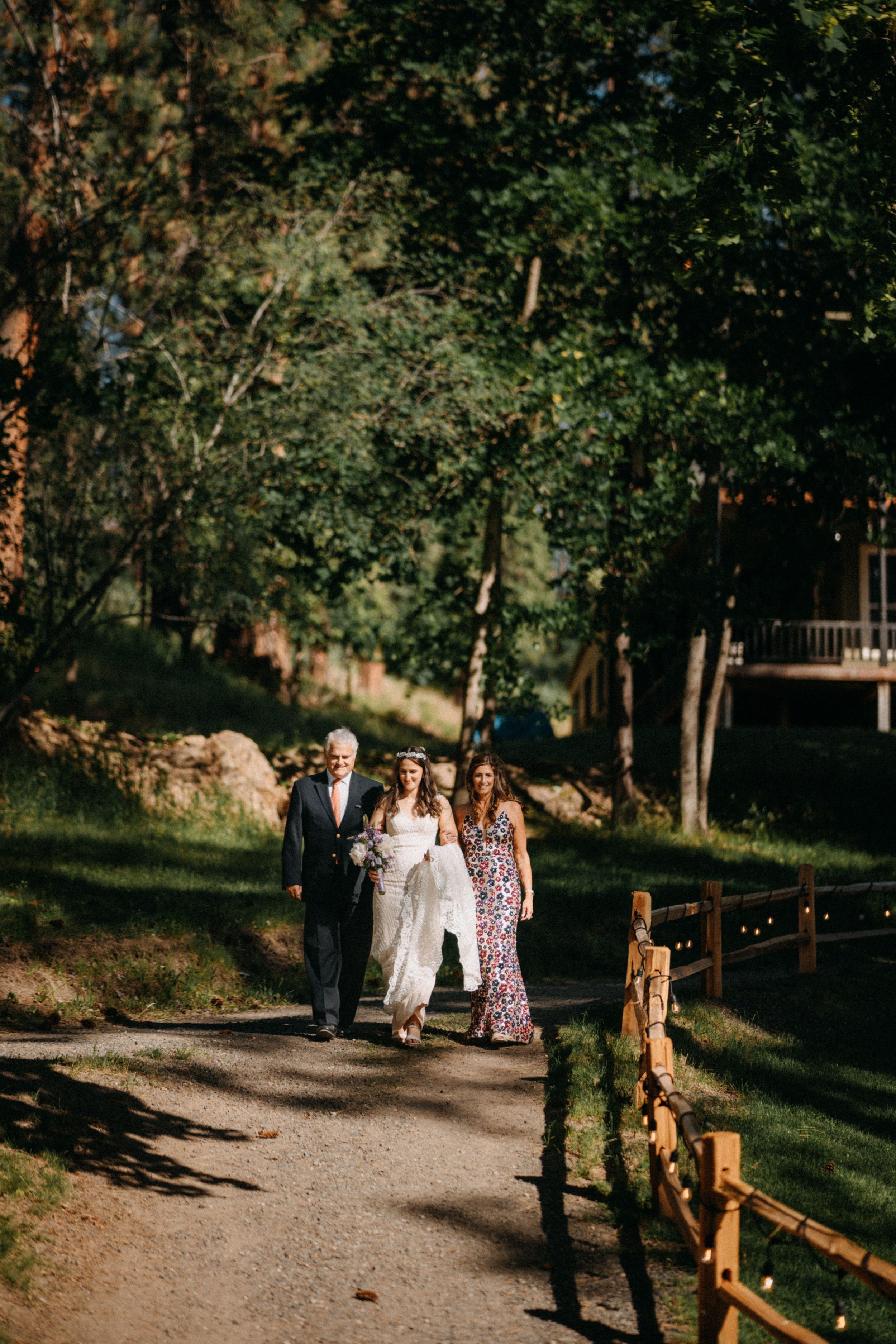 Guests walk along a rustic wooden fence-lined path through a wooded outdoor wedding venue.