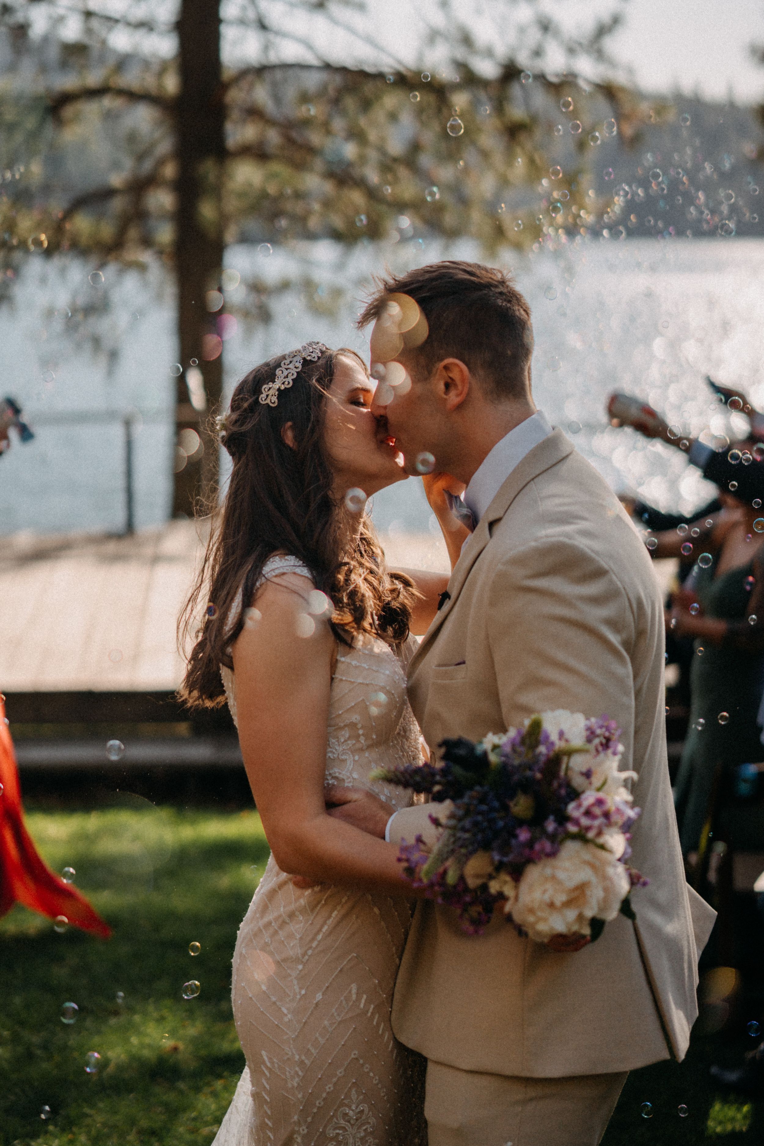 Newlyweds share a romantic kiss outdoors with bubbles floating around them.