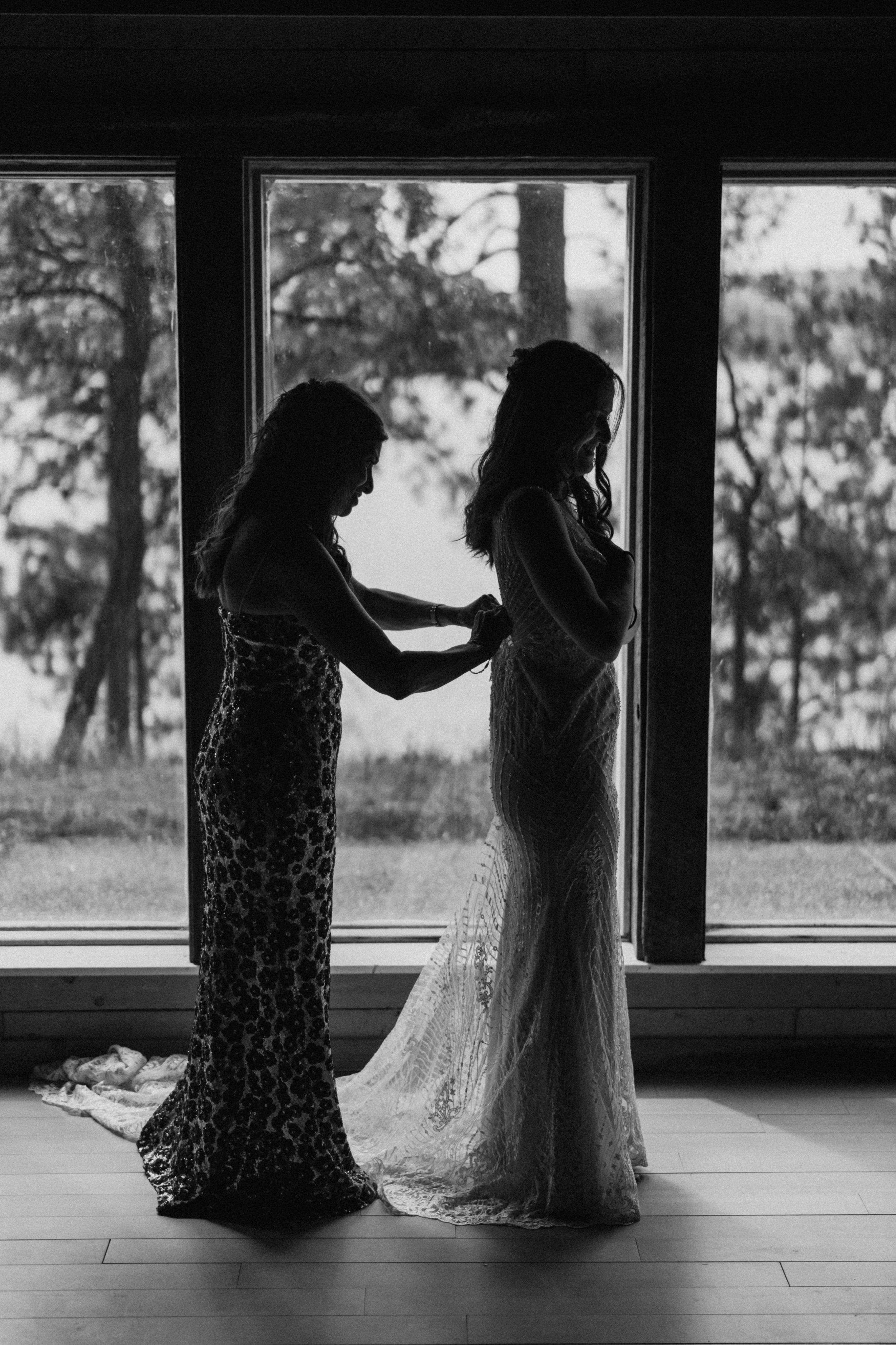 A silhouette of two figures in formal dresses against a window, one helping the other with final wedding preparations.