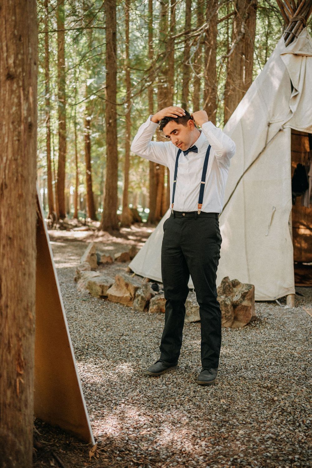 Groom in black pants and suspenders walking on gravel path through forest setting.