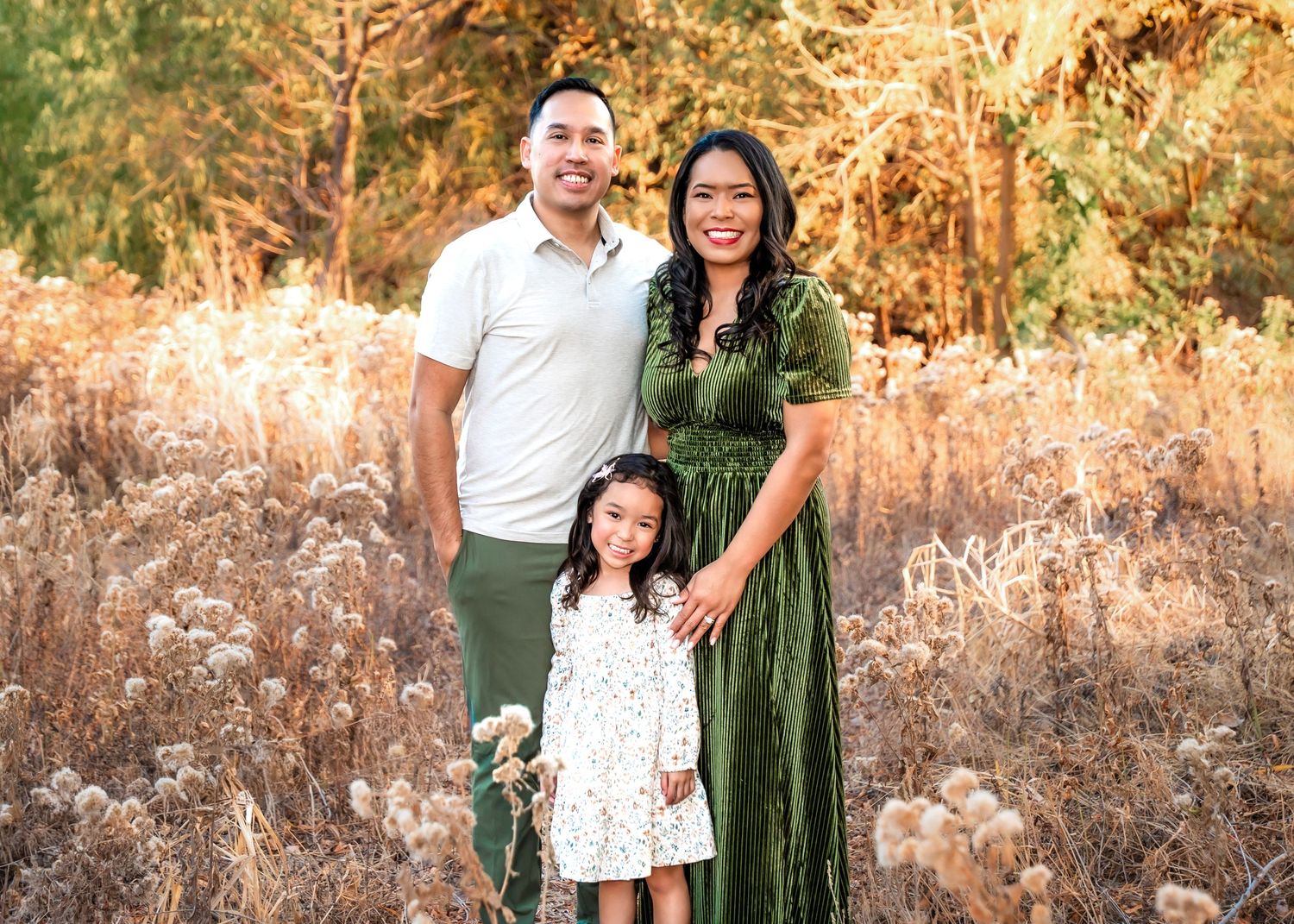 A family poses together in a natural outdoor setting with golden sunlight filtering through trees in the background.