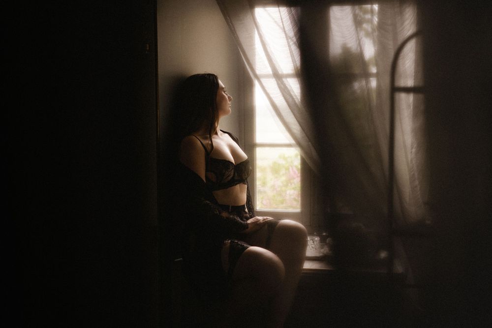 A woman in dark lingerie sits by a dimly lit window with sheer curtains in a moody boudoir setting.