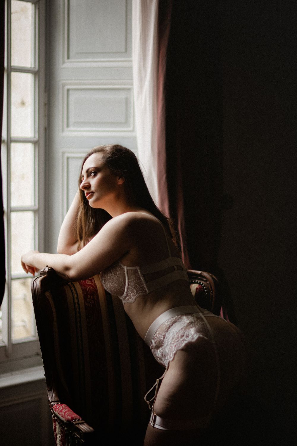 Woman in white lace lingerie sits in an ornate chair gazing out a large window with pink curtains.