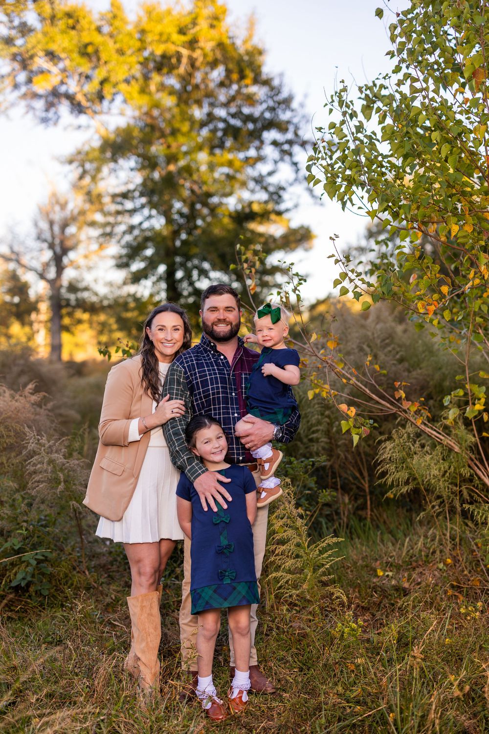 Family of four poses together in an autumn field with golden sunlight filtering through trees.