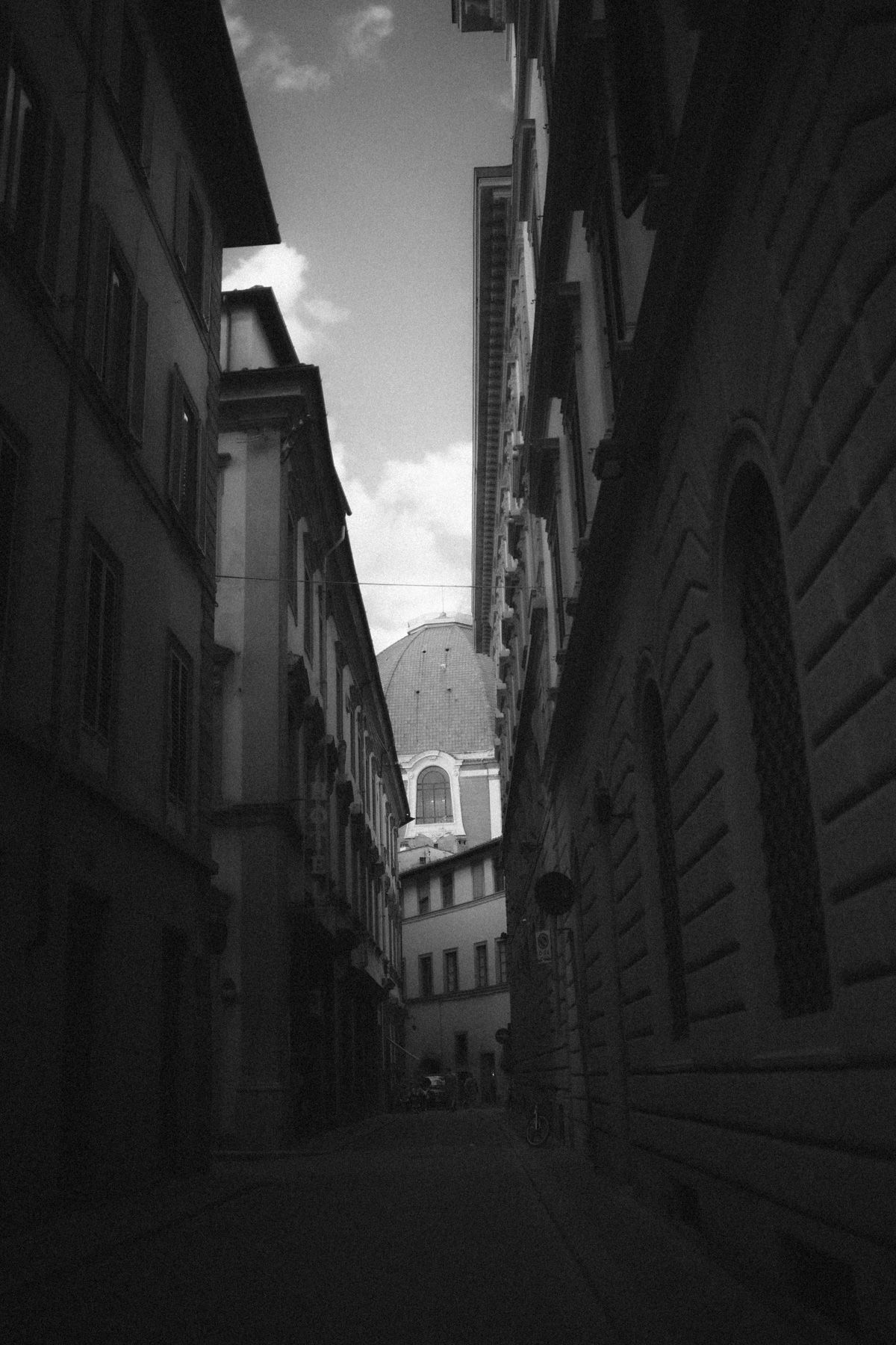 A narrow black and white street view in Florence, Italy with historic buildings leading to a domed church in the distance.