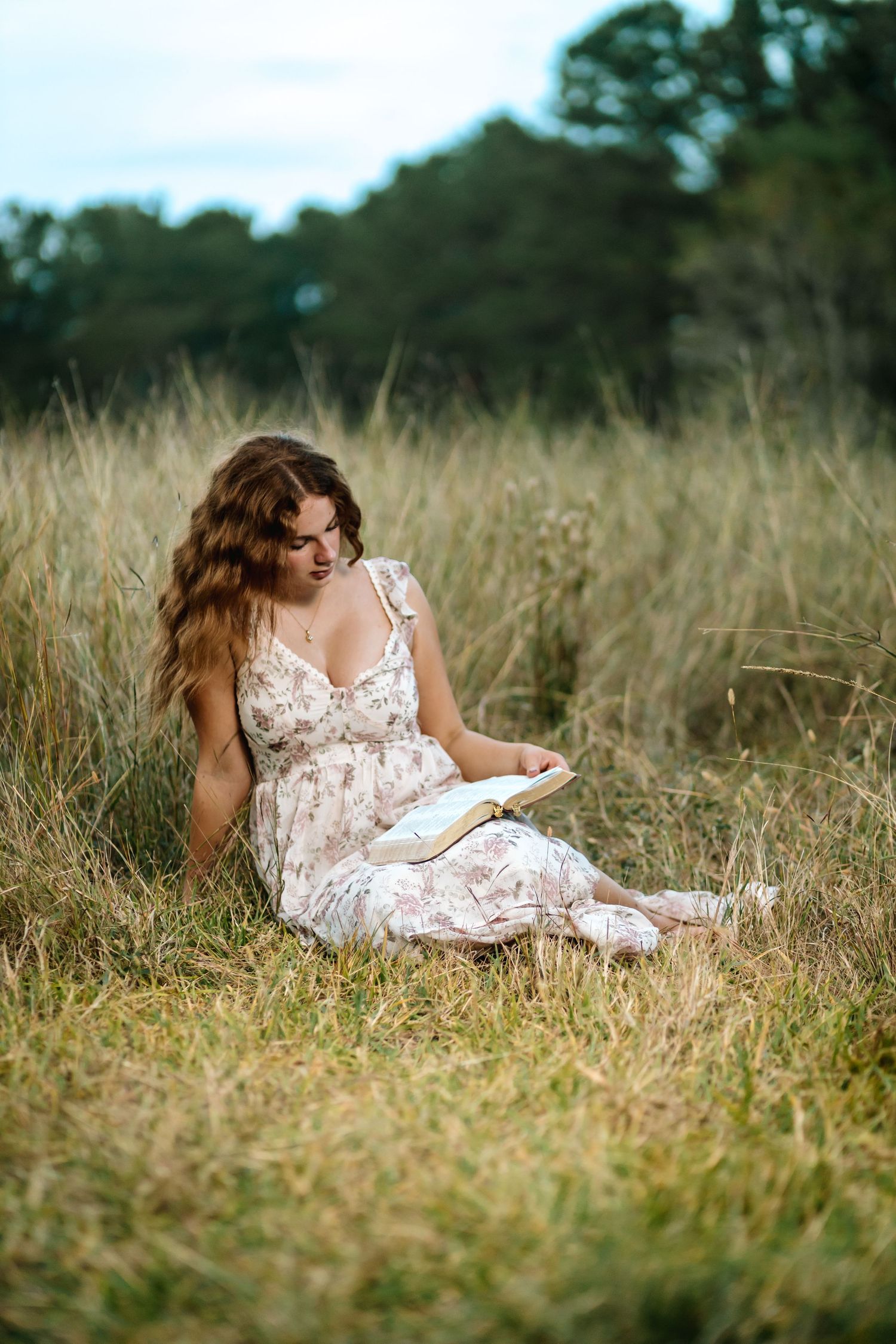 A person in a white floral dress reclines in a sun-dappled meadow of tall grass while reading a book.