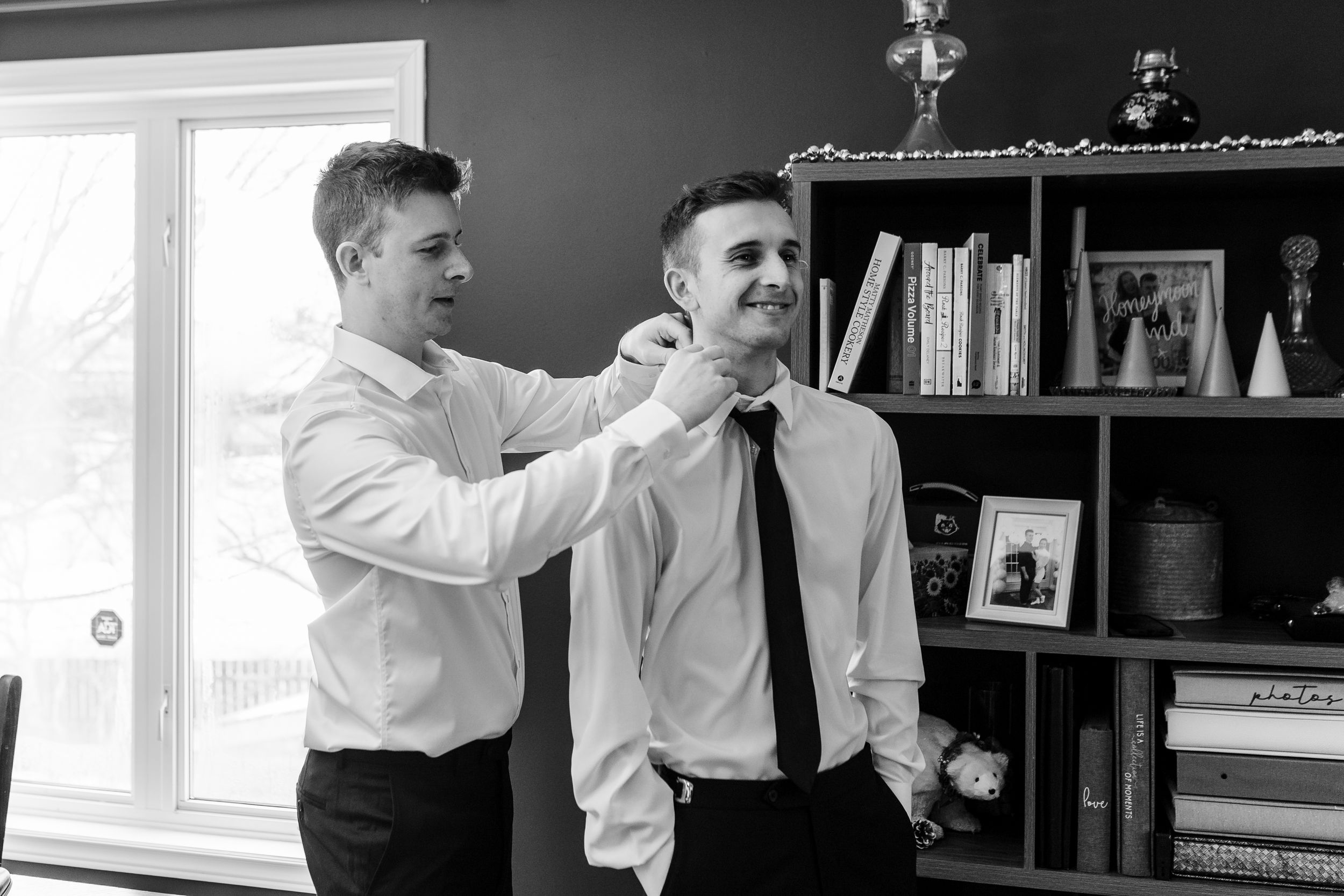 Two men in white dress shirts and black ties stand together while one adjusts the other's tie in a casual office setting.