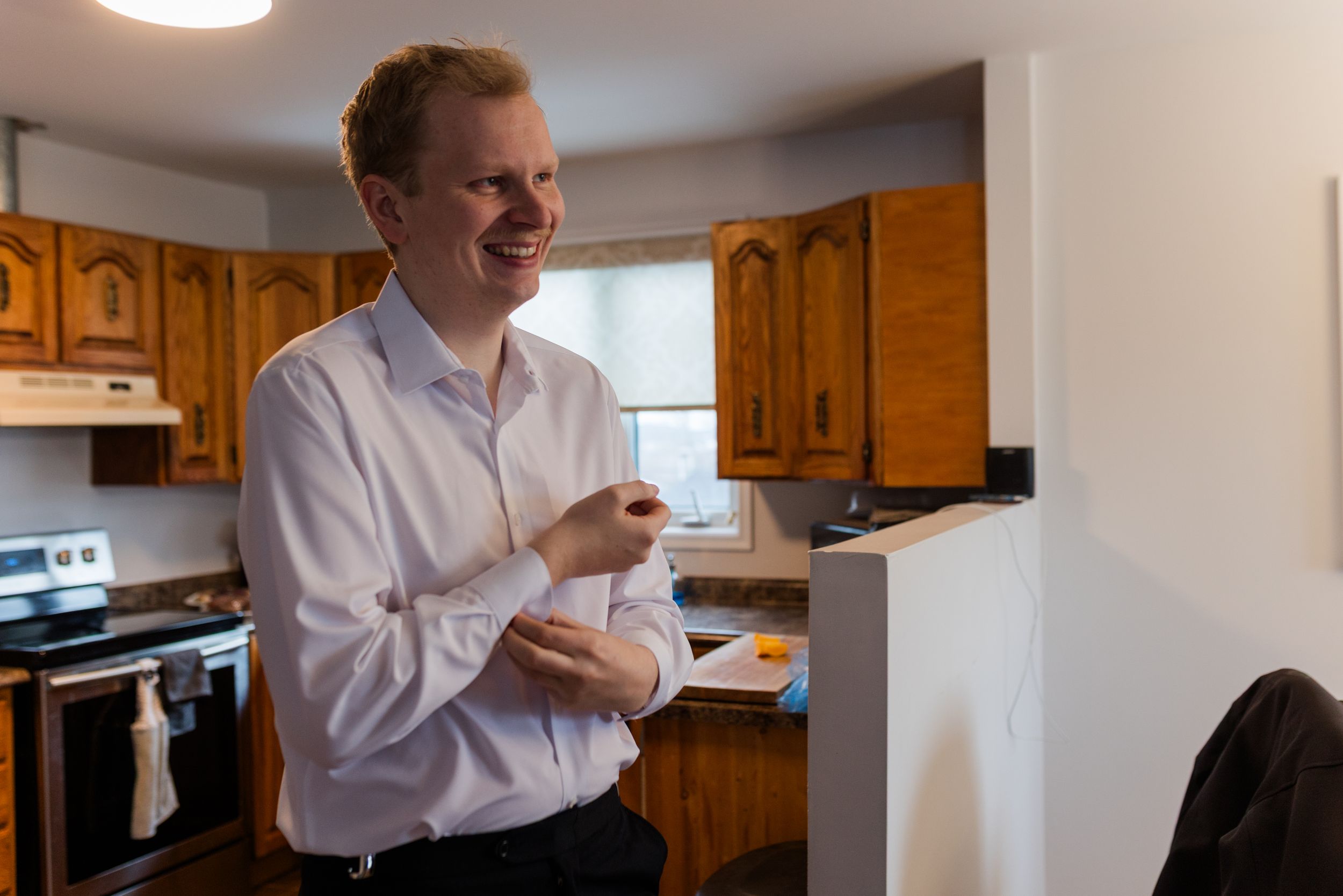 Person in white button-down shirt standing in kitchen with wooden cabinets and natural lighting.