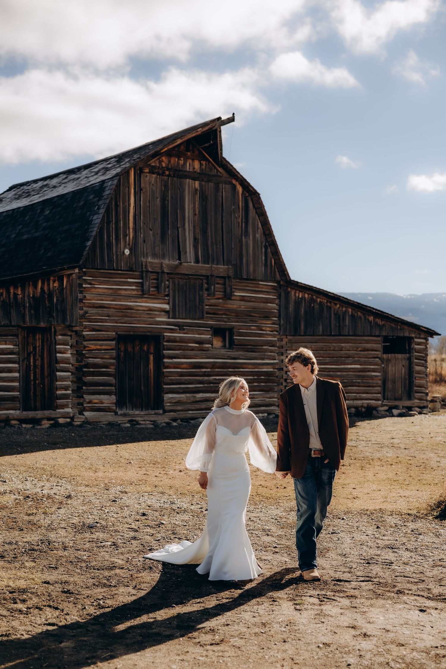 A couple walks through a rustic setting with weathered wooden barns against a cloudy sky.