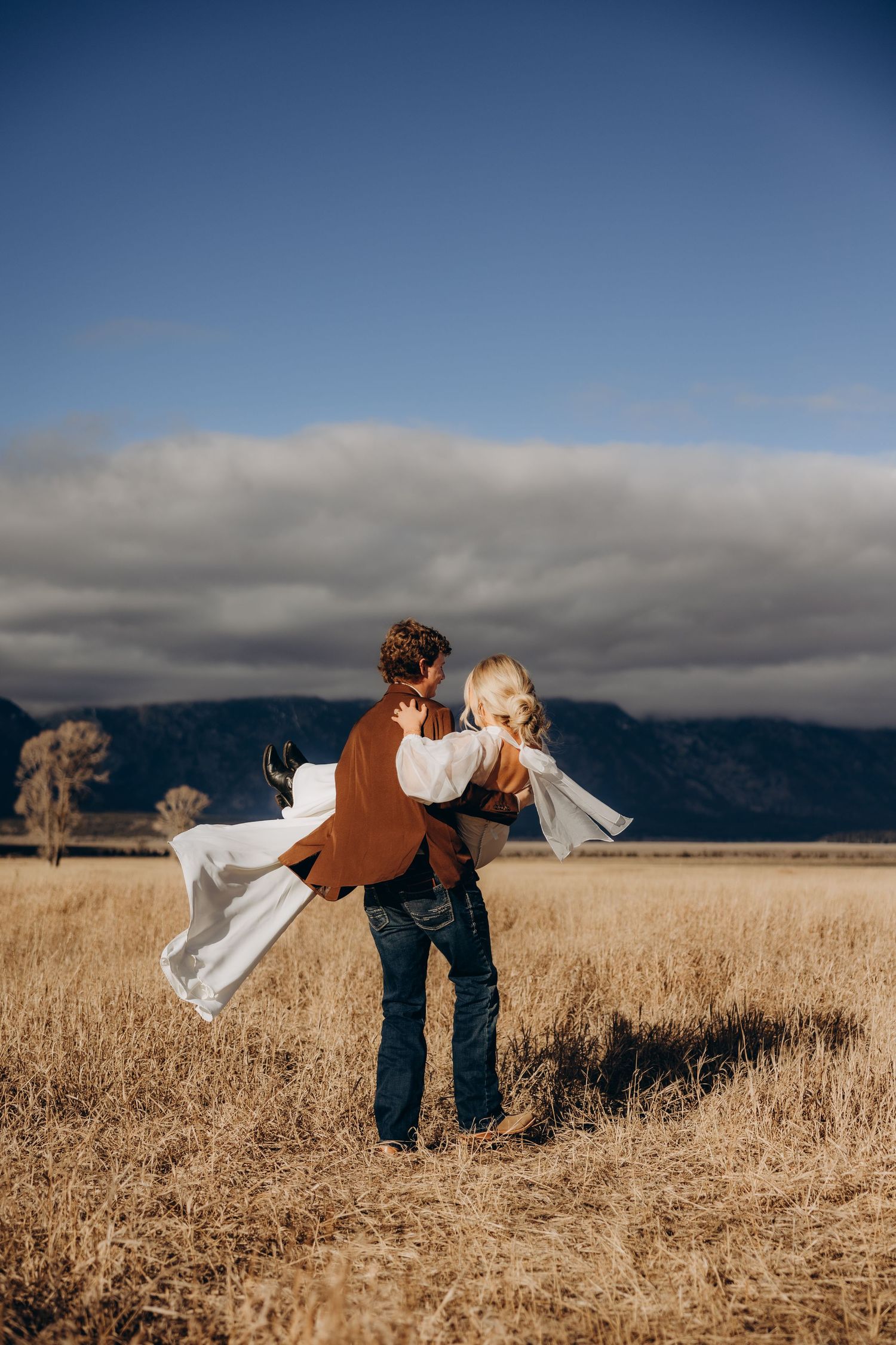 A couple dances playfully in a golden wheat field against a dramatic cloudy sky and mountain backdrop.