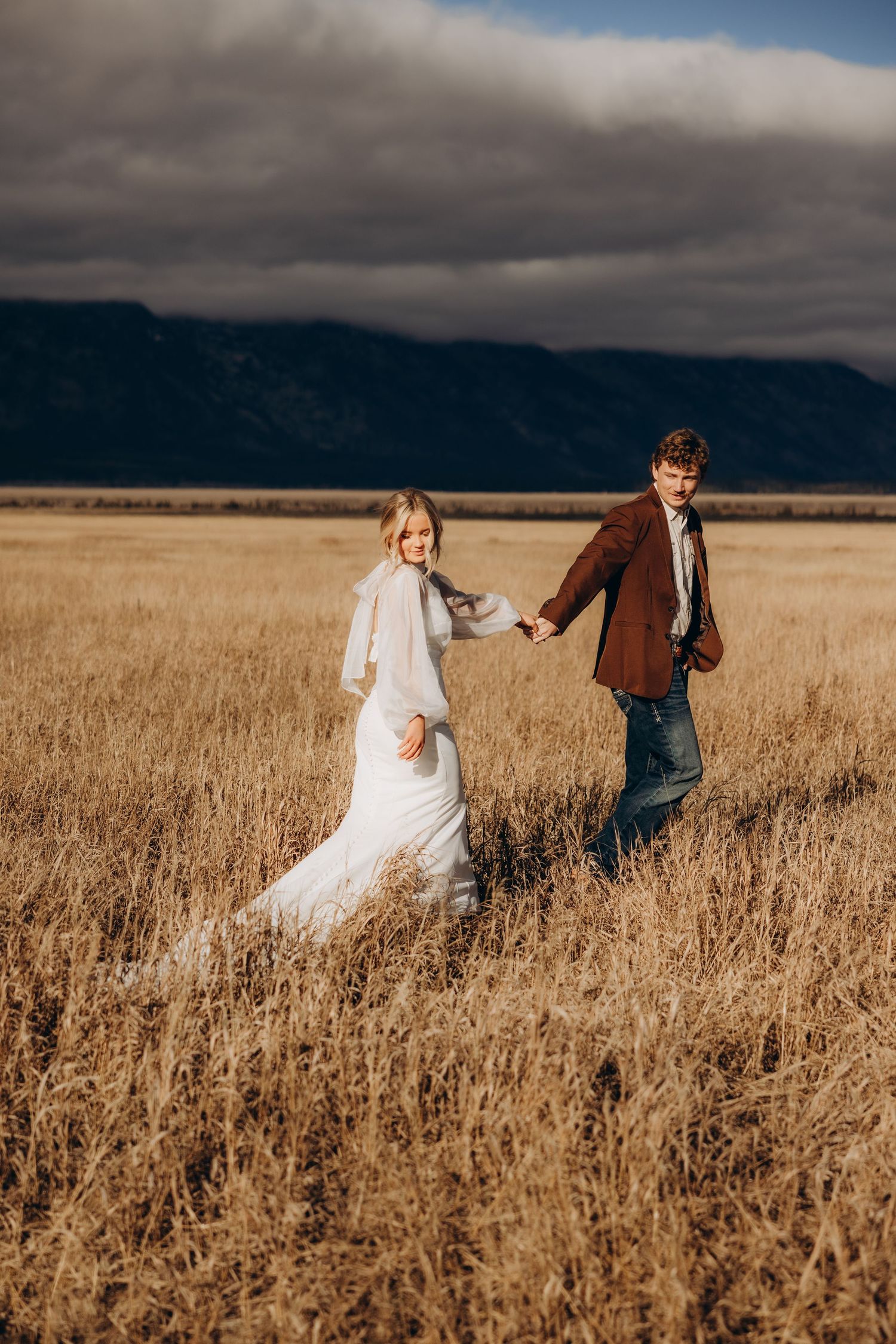 A couple runs playfully through a golden wheat field under dramatic storm clouds.