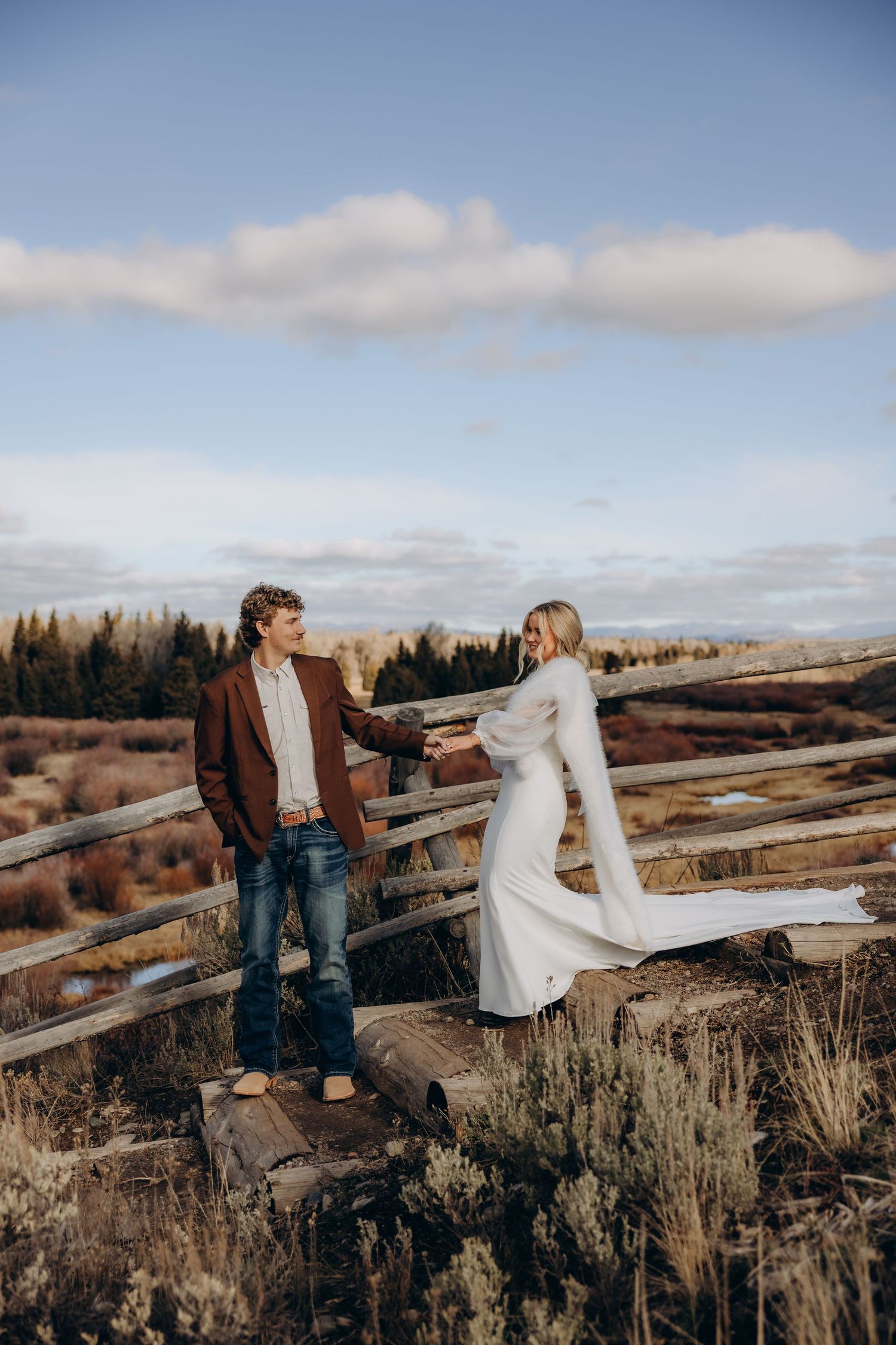Couple playfully interacting on a wooden fence with mountains and forest in the background.