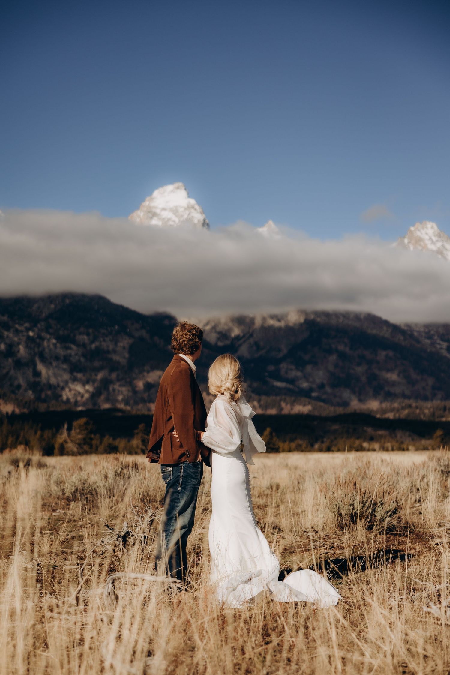 Couple stands in golden field with dramatic mountain landscape and cloudy sky behind them.