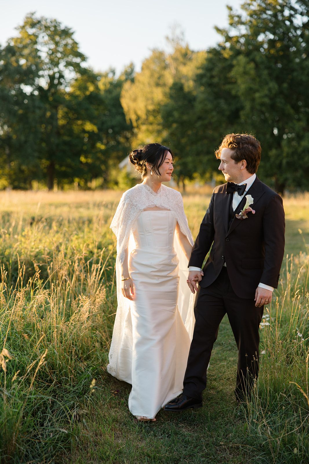 A couple in wedding attire walks together through a sunlit meadow with tall grass and trees in the background.