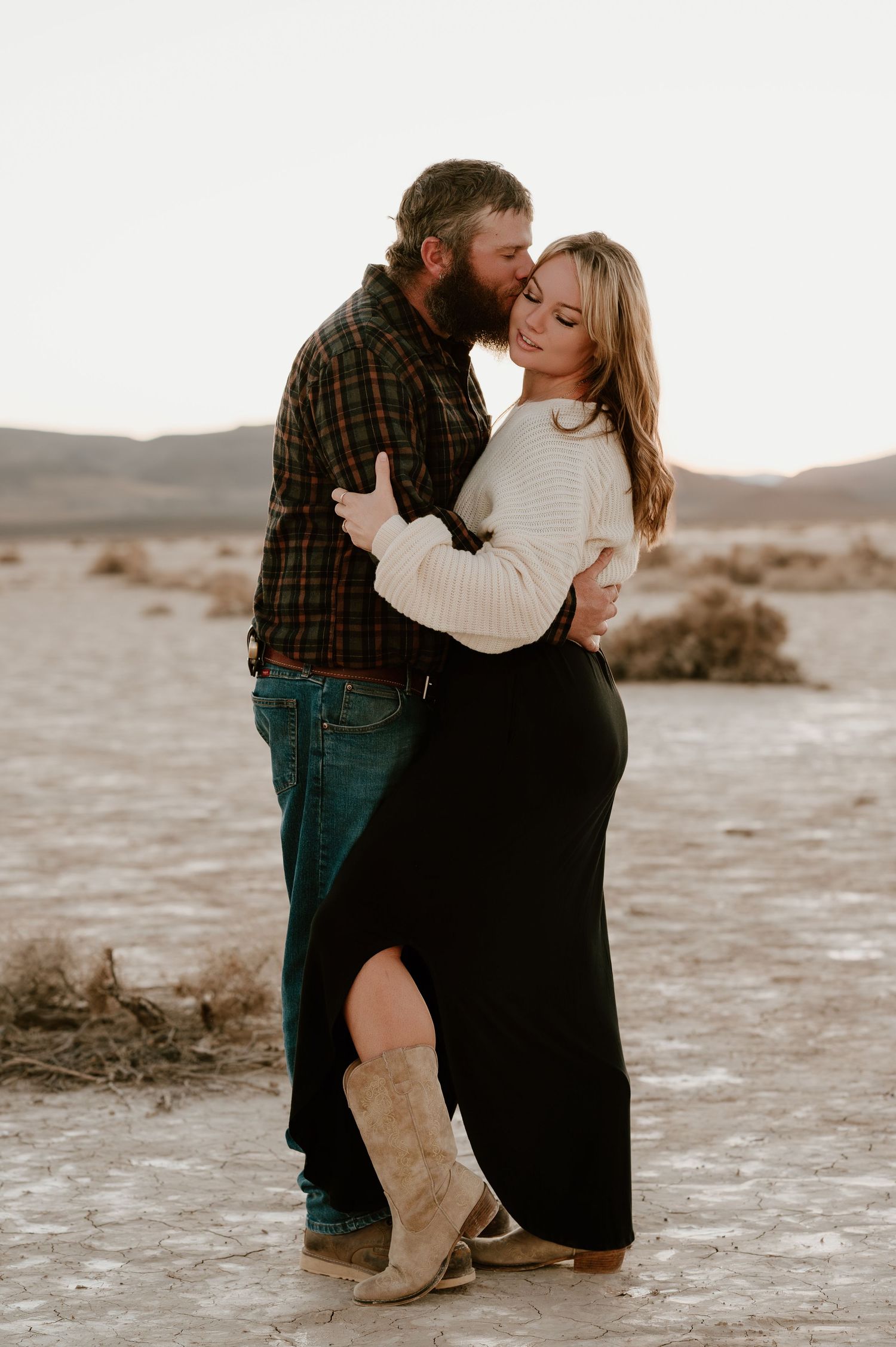 A romantic couple embraces in the desert, with her wearing a cream sweater and black maxi skirt with cowboy boots.