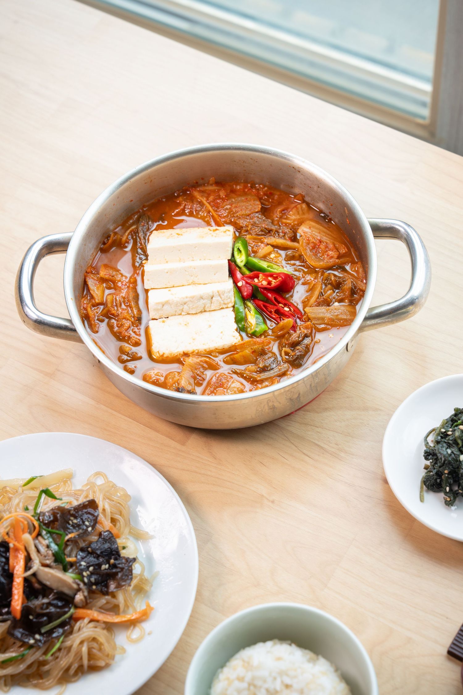 Korean food dishes including kimchi stew, rice, and side dishes served in traditional bowls on a wooden table.