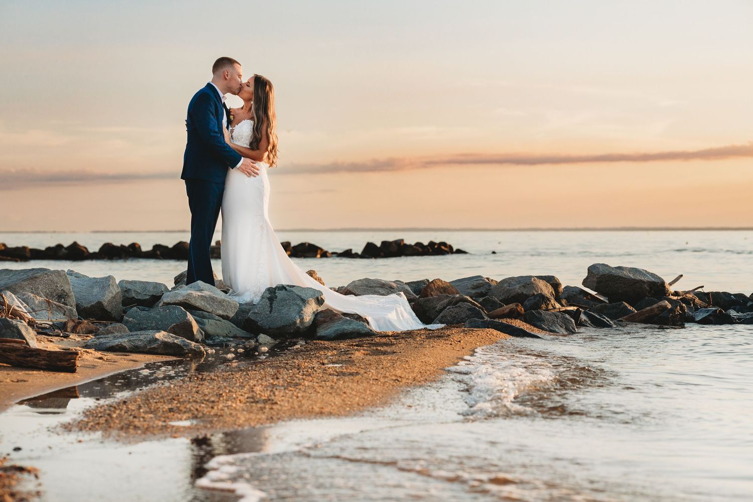 Sunset beach wedding photos with couple silhouetted against golden sky and rocky shoreline in Kent Island Maryland. Annapolis videographer