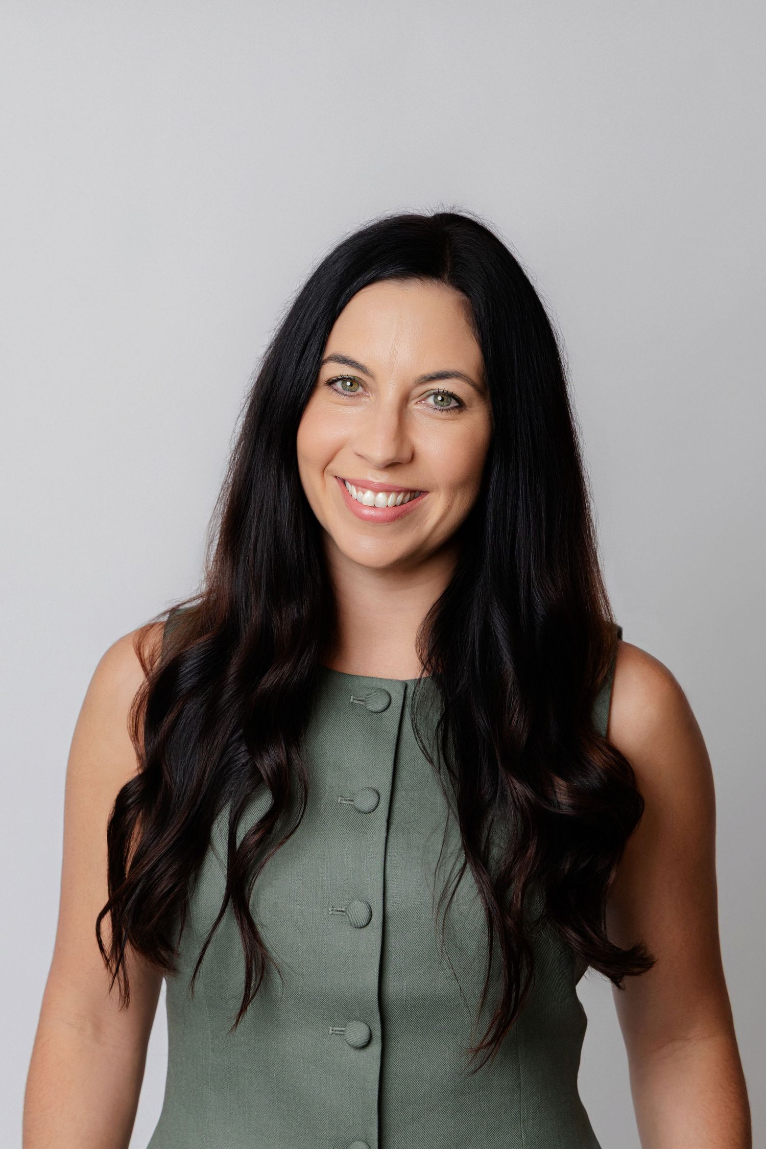 Professional headshot of a woman with long dark hair wearing a sage green sleeveless button-up top against a gray background.