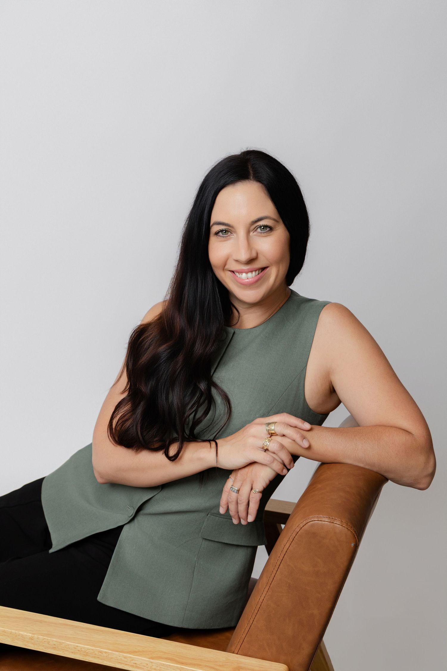 Woman in sage green sleeveless top seated on brown leather chair against neutral background.