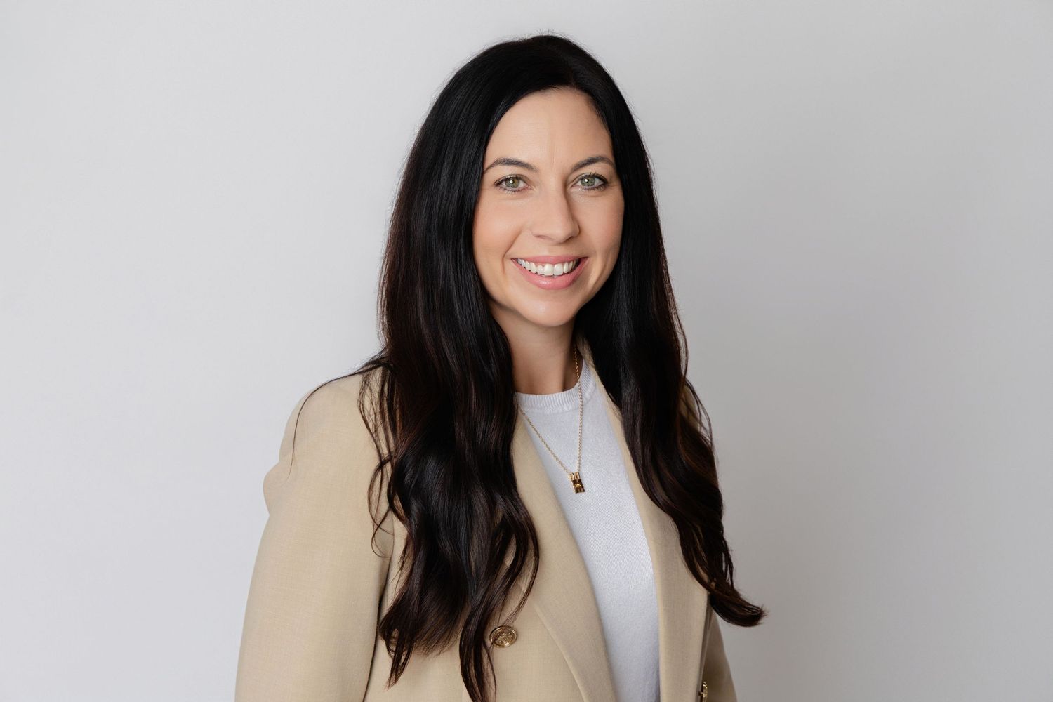 Professional headshot of someone with dark hair wearing a white top and beige blazer against a light gray background.