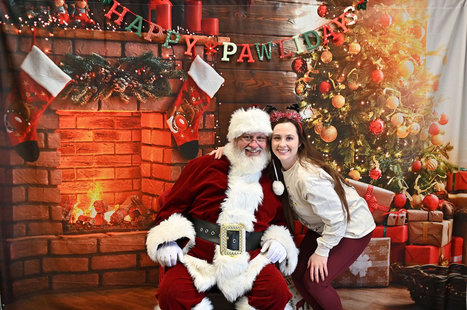 Santa Claus poses with pets at a festive holiday photo session against a red brick wall and Christmas decorations.
