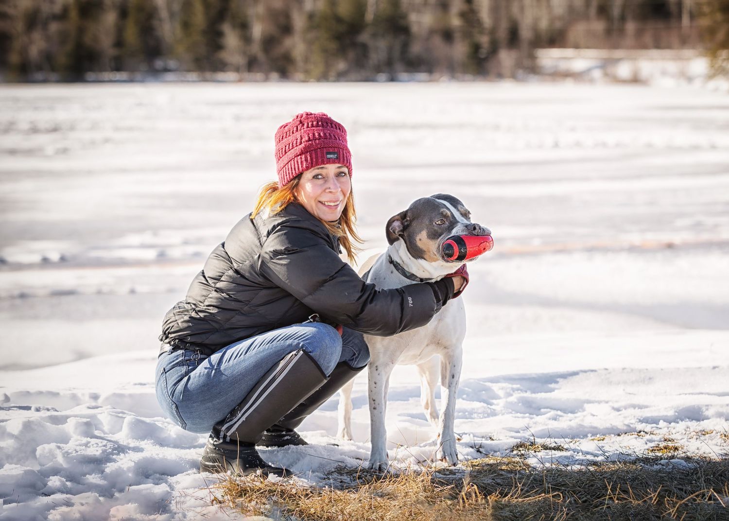 A person in a red knit hat and winter coat crouches next to a gray dog wearing a red bandana in snowy landscape.