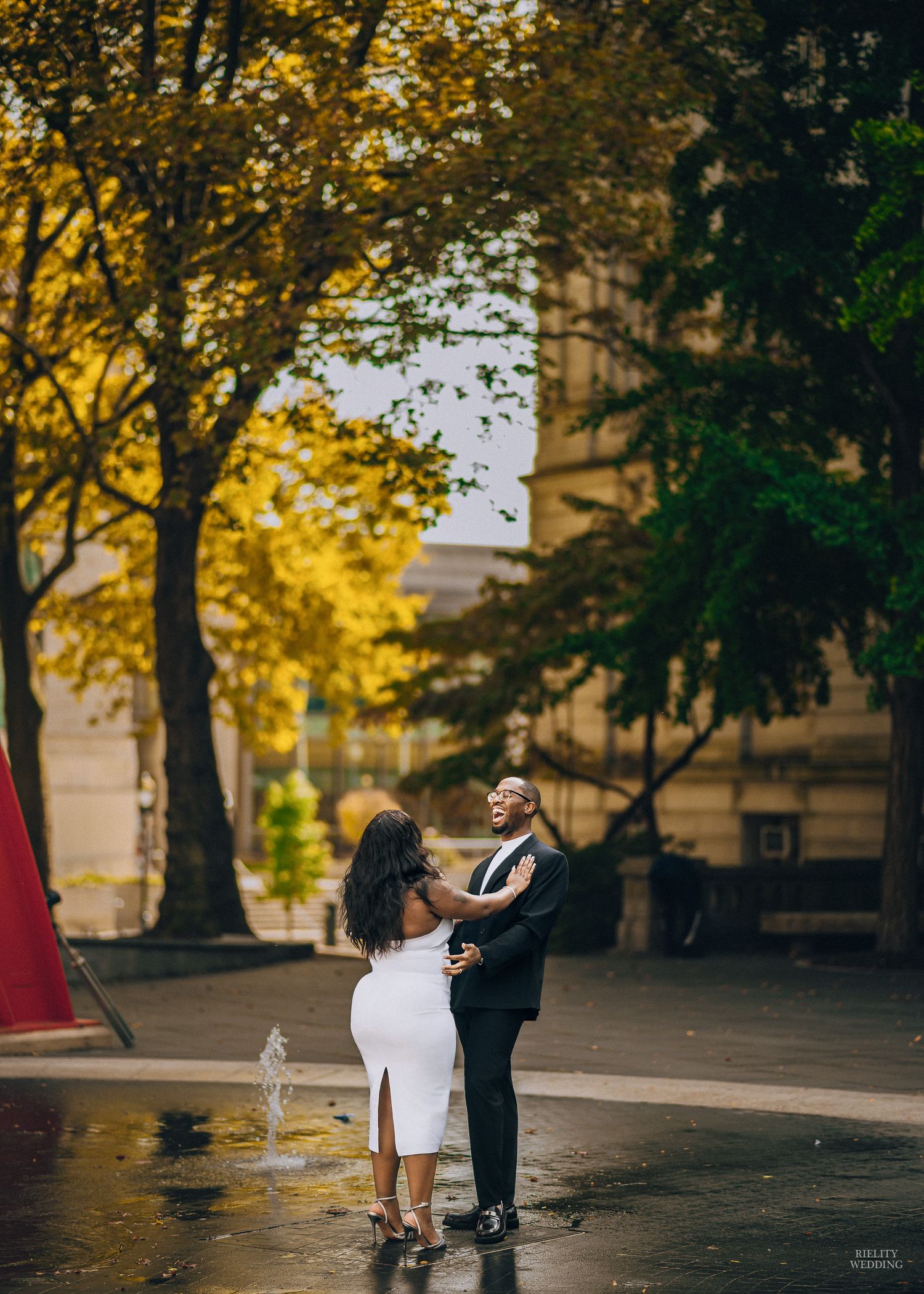 A romantic couple dances together in a golden autumn setting with sunlit trees and fallen leaves on the ground.