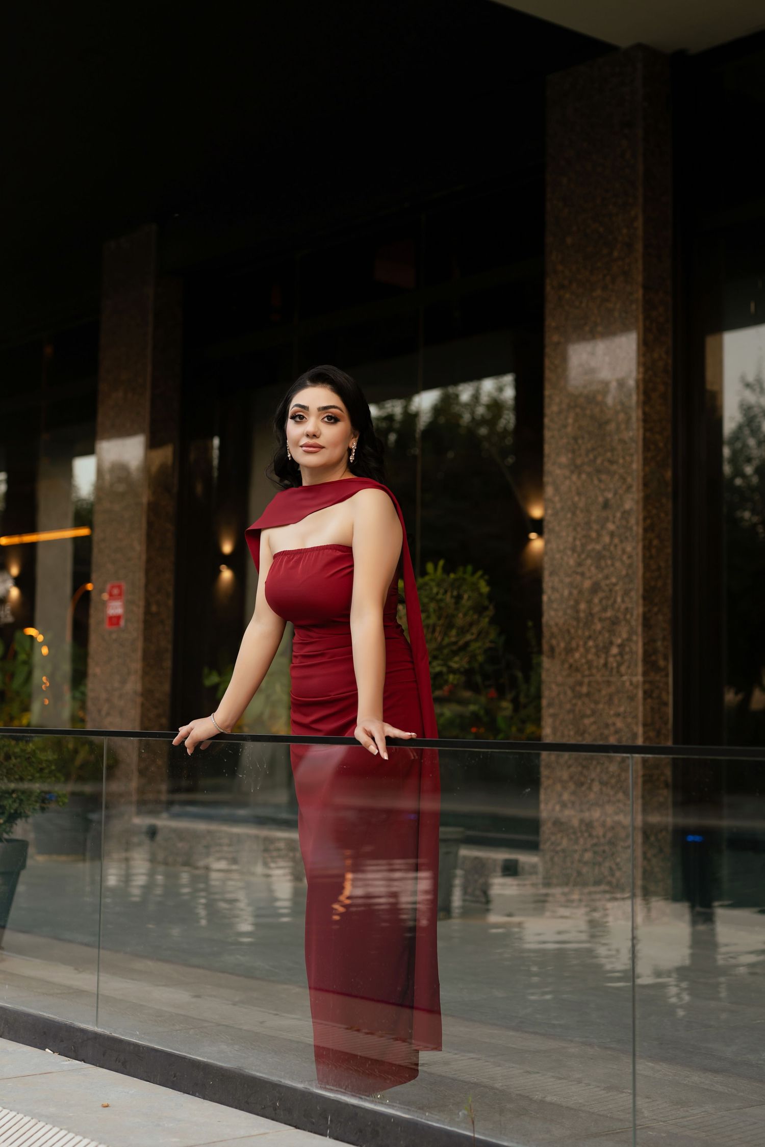 A person in a burgundy dress poses elegantly against glass railings in a modern architectural setting.