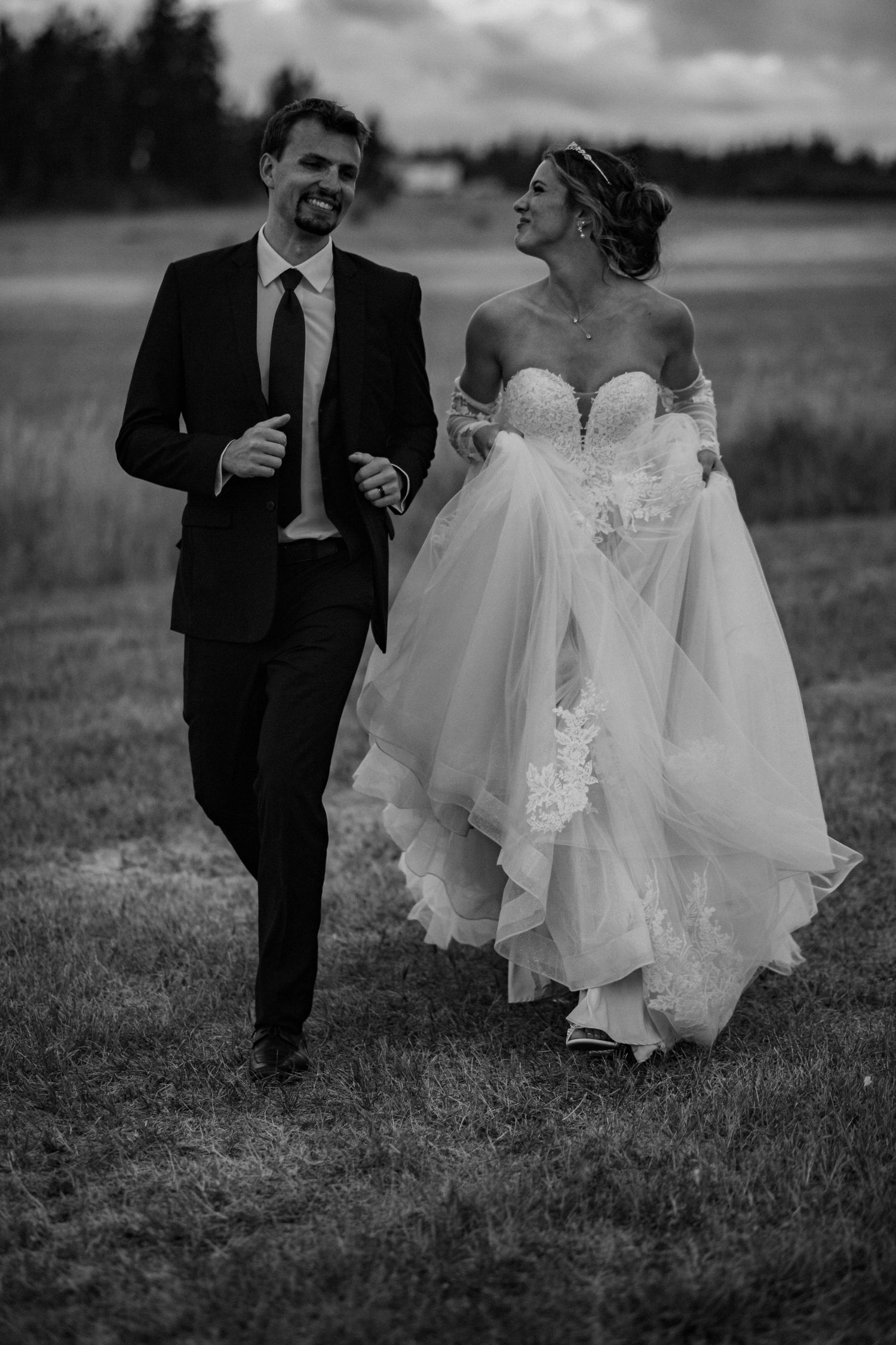A black and white wedding photo showing a couple standing in a field at dusk with blurred foliage in the foreground.