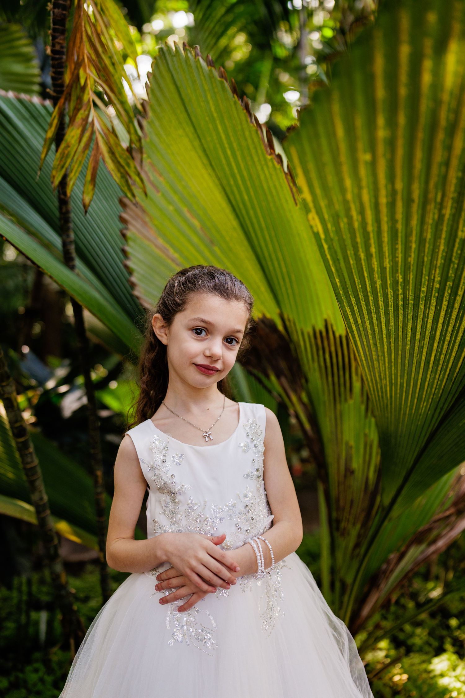 A white formal dress with lace details stands out against lush tropical palm fronds in a garden setting.