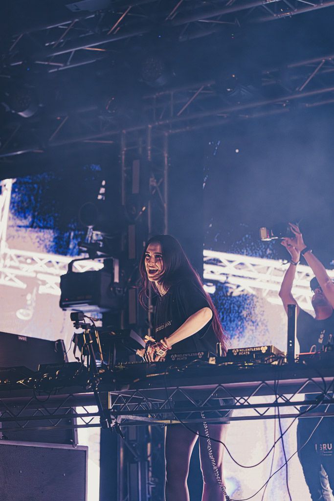 Concert performers energetically operate electronic music equipment on stage with dramatic blue lighting and fog effects.